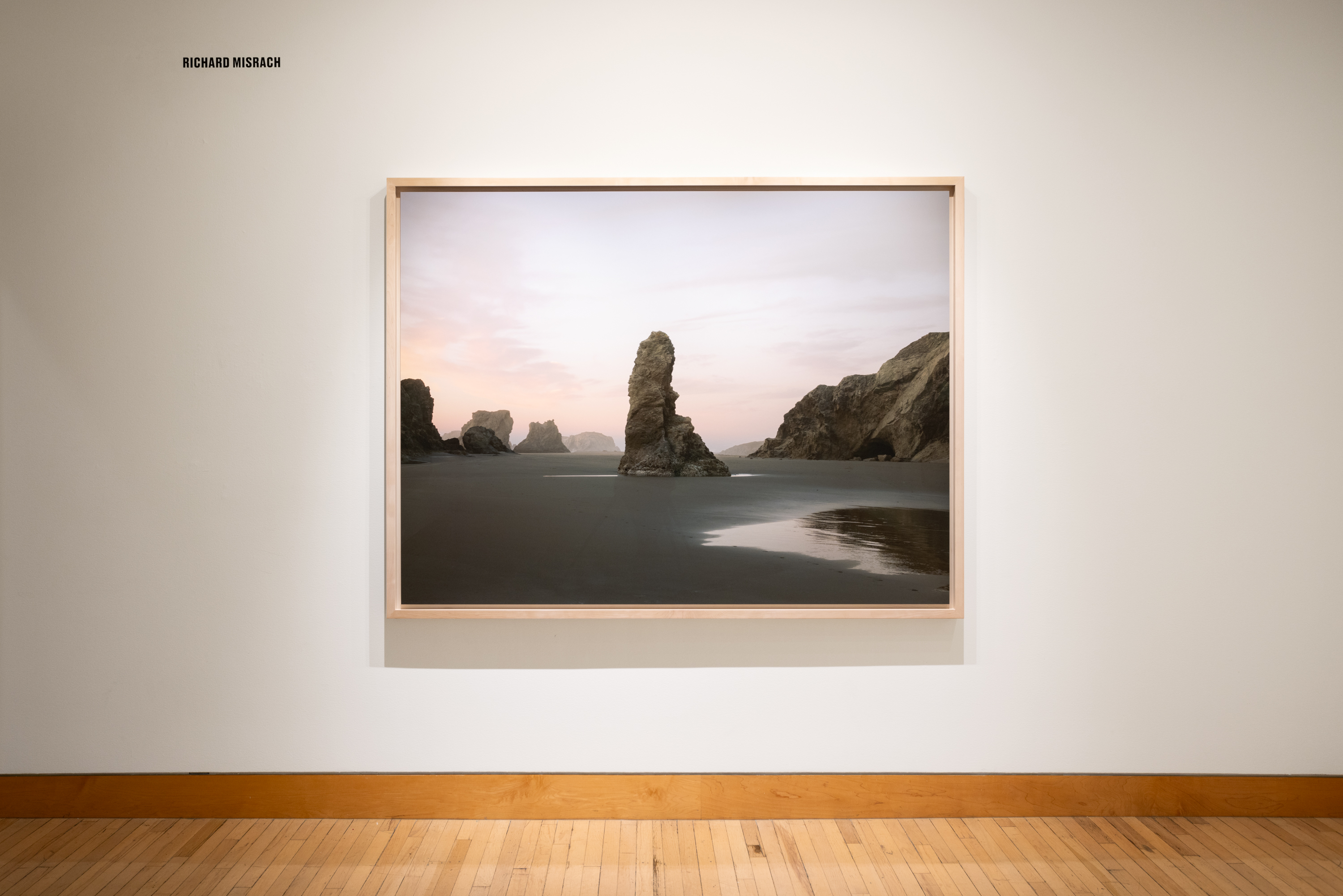 Color image of color photograph depicting a larger rock formation on the beach framed in bleached wood on white gallery wall