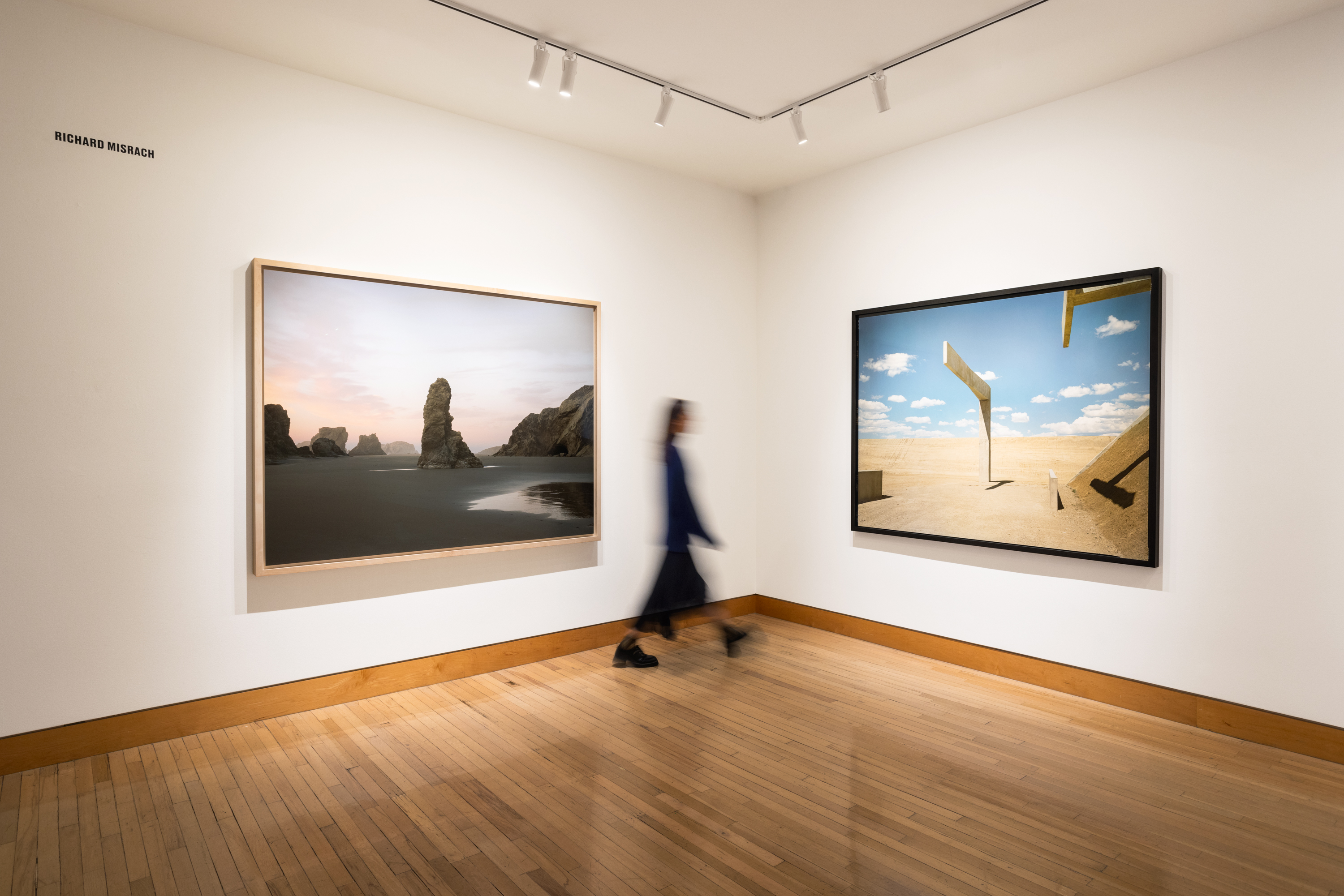 Color image of two color photographs depicting a coastal landscape and a desert landscape framed in bleached wood and black on white gallery walls