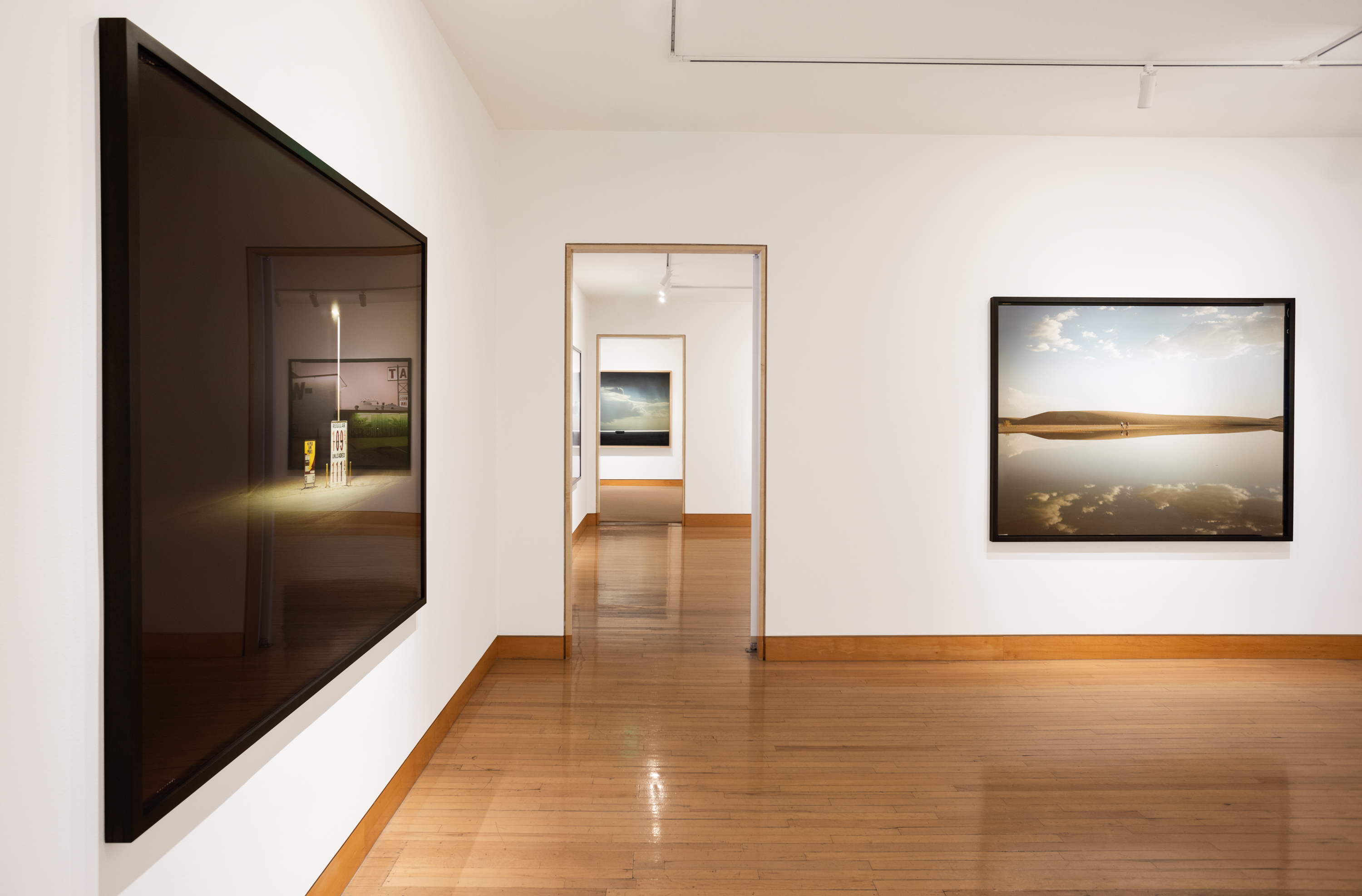 Color image of an exhibition of color photographs depicting various landscapes in the desert and by water on white gallery walls