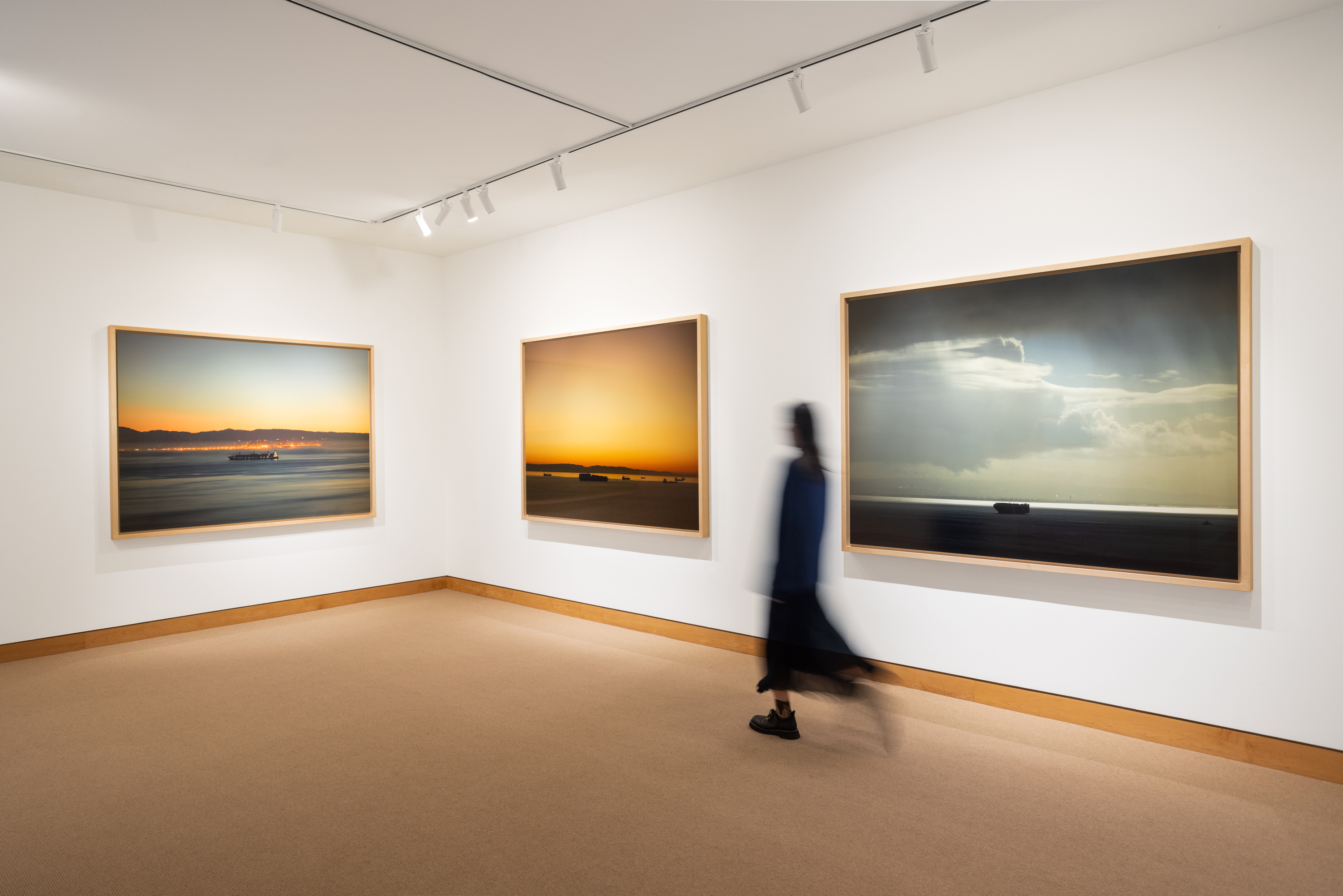 Color image of three color photographs depicting cargo ships on water at different times of day framed in bleached wood on white gallery walls