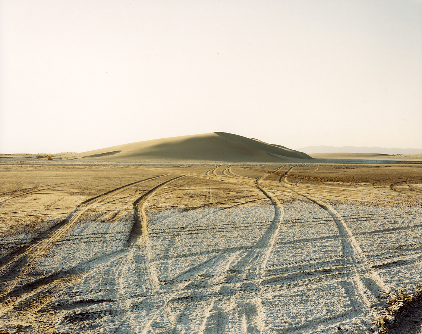 Color photograph of car tracks on desert ground with a sand hill on the horizon
