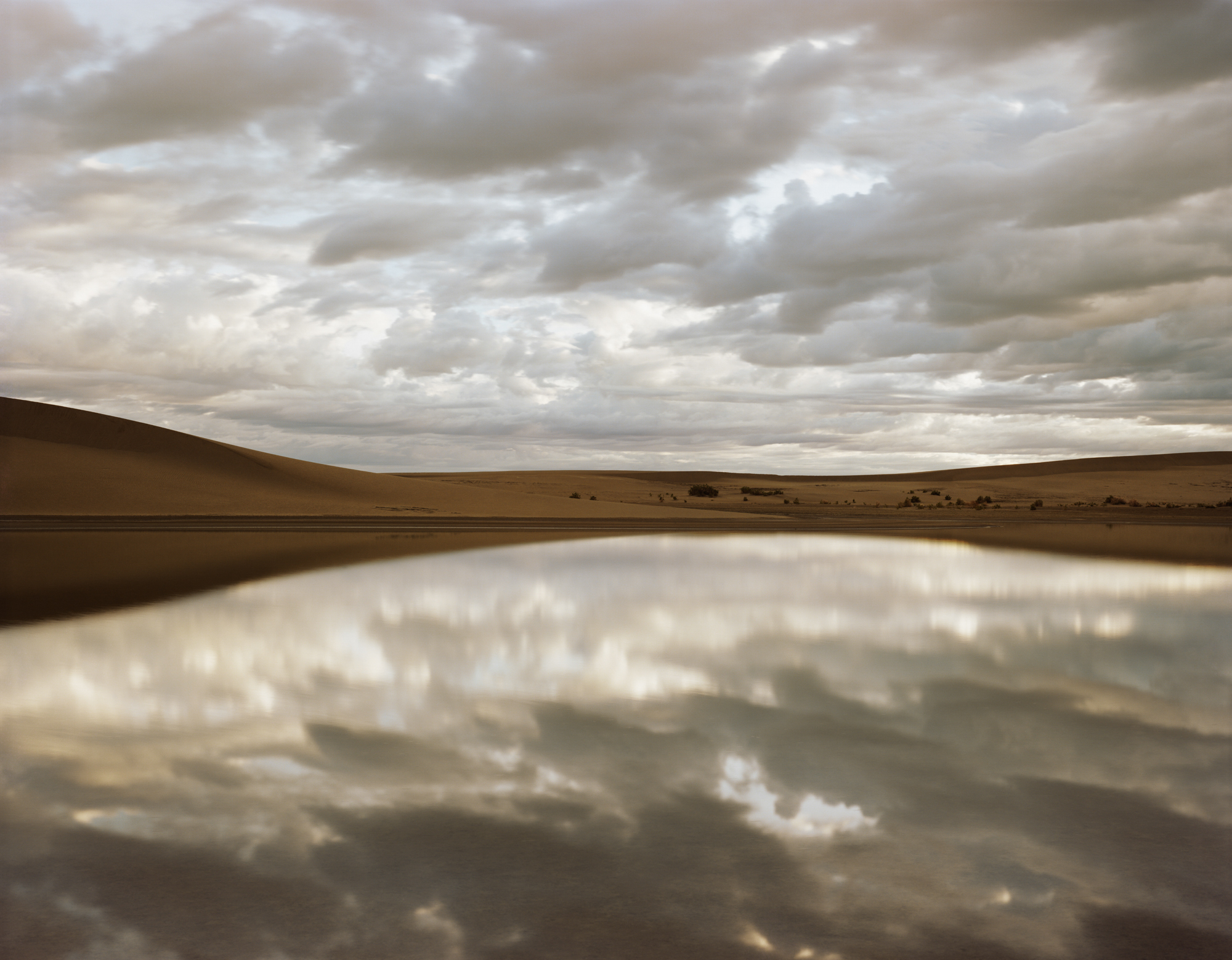 Color photograph of a lake within a desert reflecting the cloudy sky above
