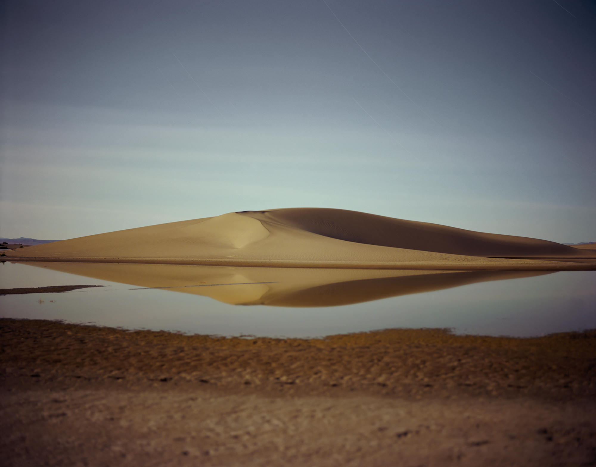Color photograph of a small body of water in front of a large sand mound with clear blue sky