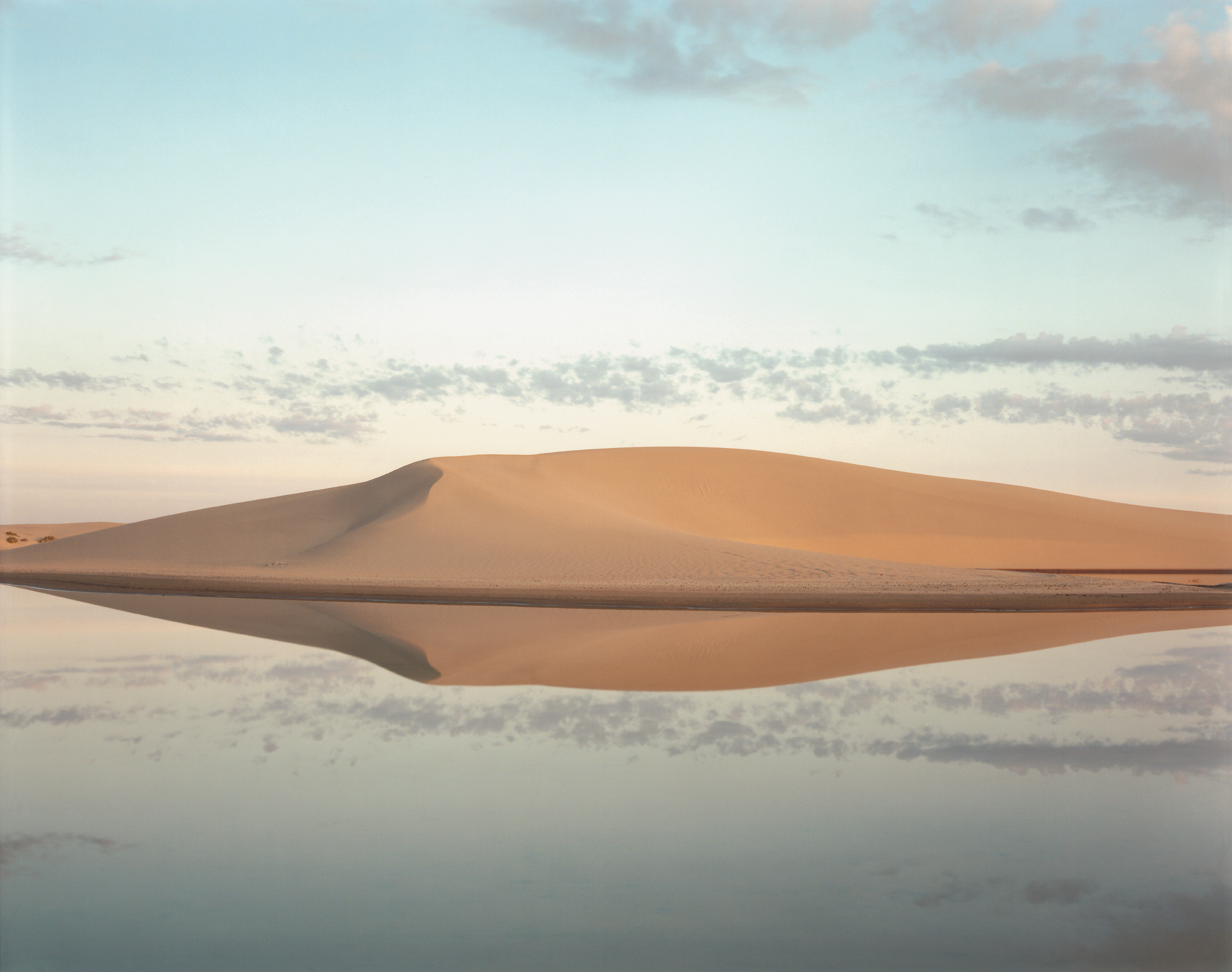 Color photograph of a small body of water in front of a large sand mound with a lightly clouded blue sky