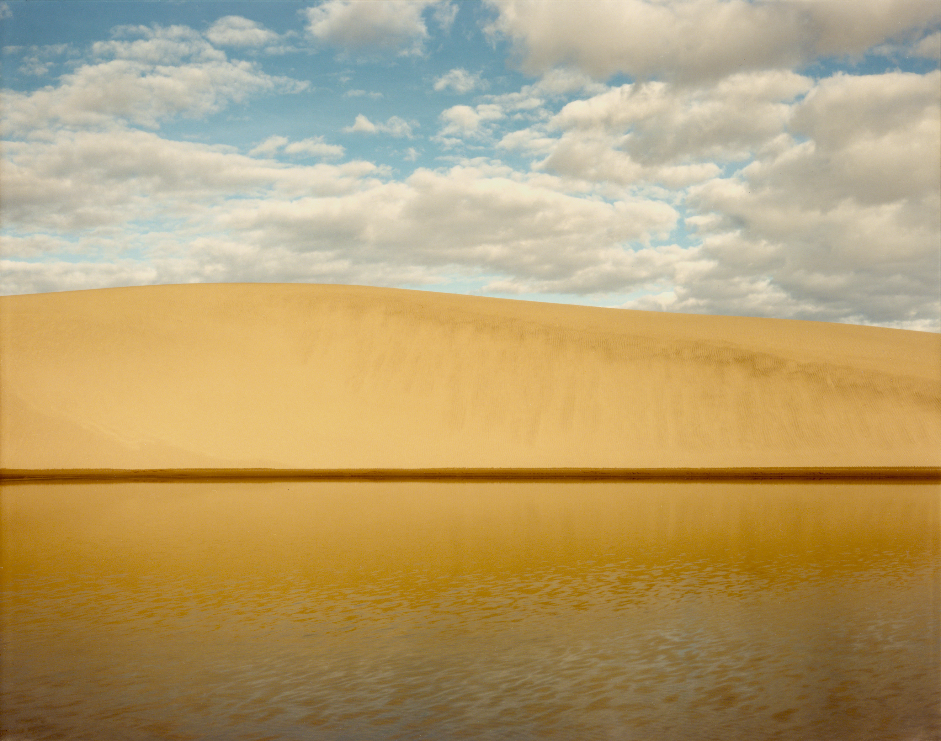 Color photograph of a body of water near a sand hill with a bright blue cloudy sky