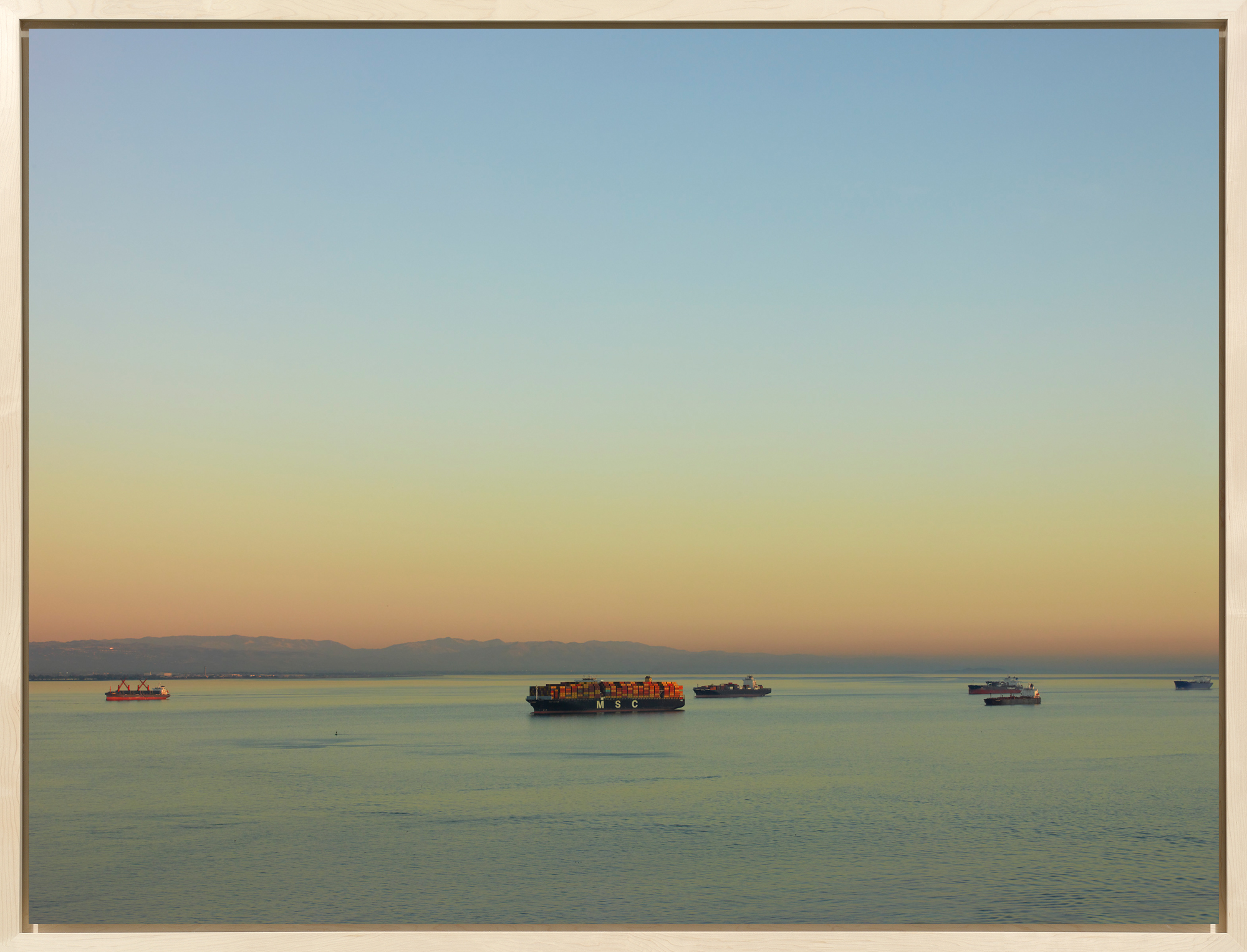 Color image of a color photograph of six cargo ships traveling along the water on a clear day framed in bleached wood