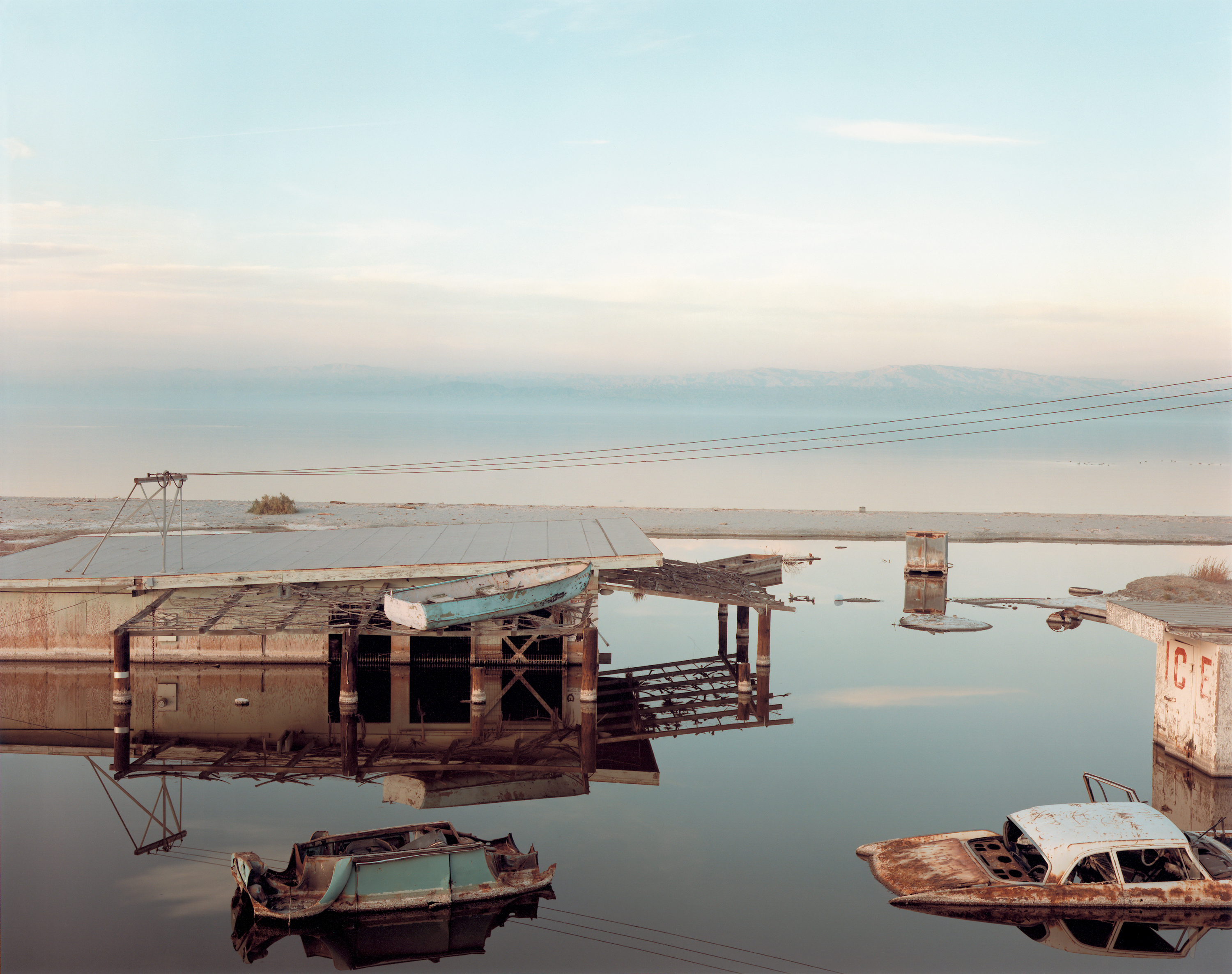 Color photograph of an abandoned building with abandoned cars and rowboats slightly submerged in water with mountain range on horizon