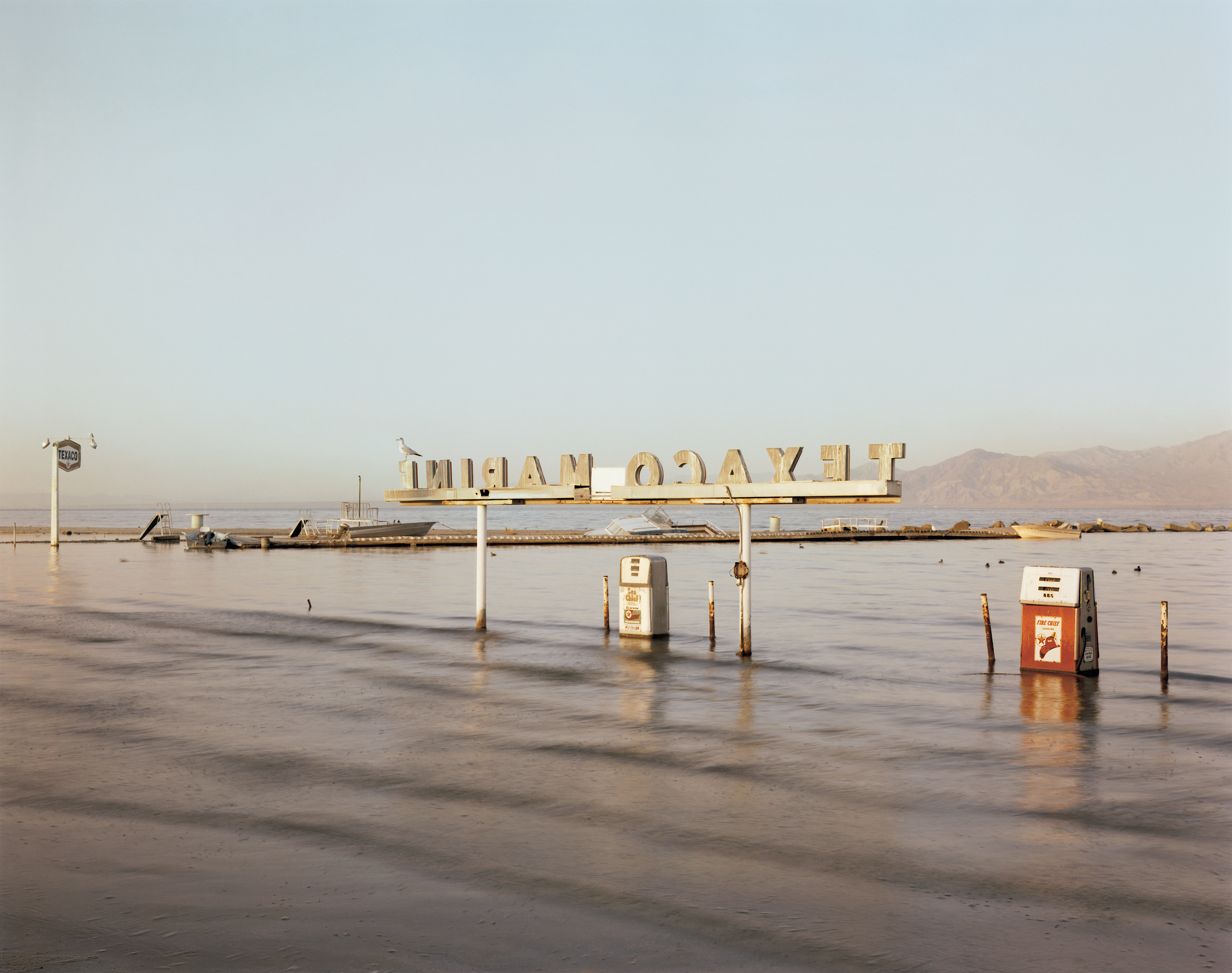 Color photograph of a submerged gas station in large body of water with mountain range on the horizon