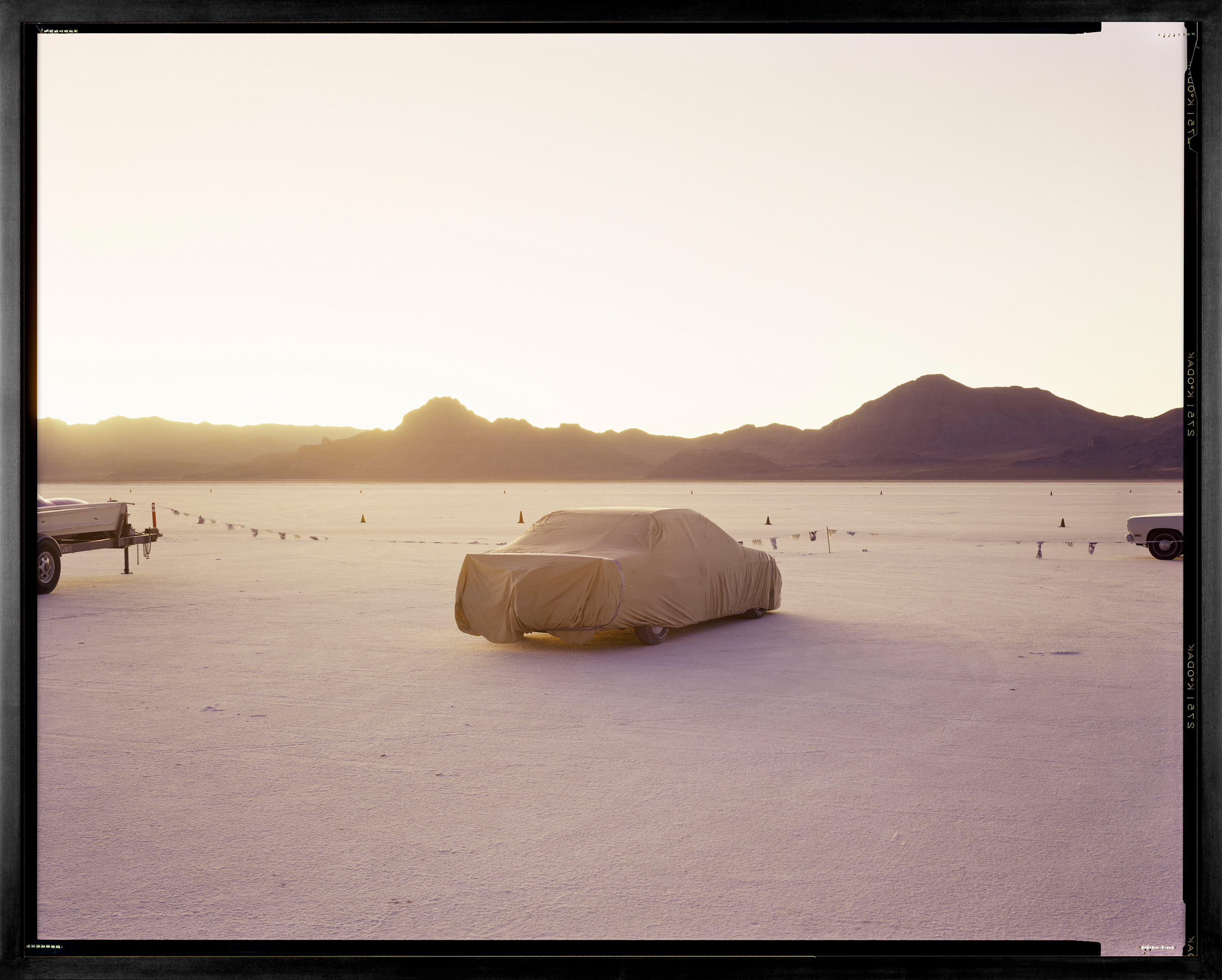 Color photograph framed in black depicting a covered car within the desert with sunlight breaking over mountains on the horizon