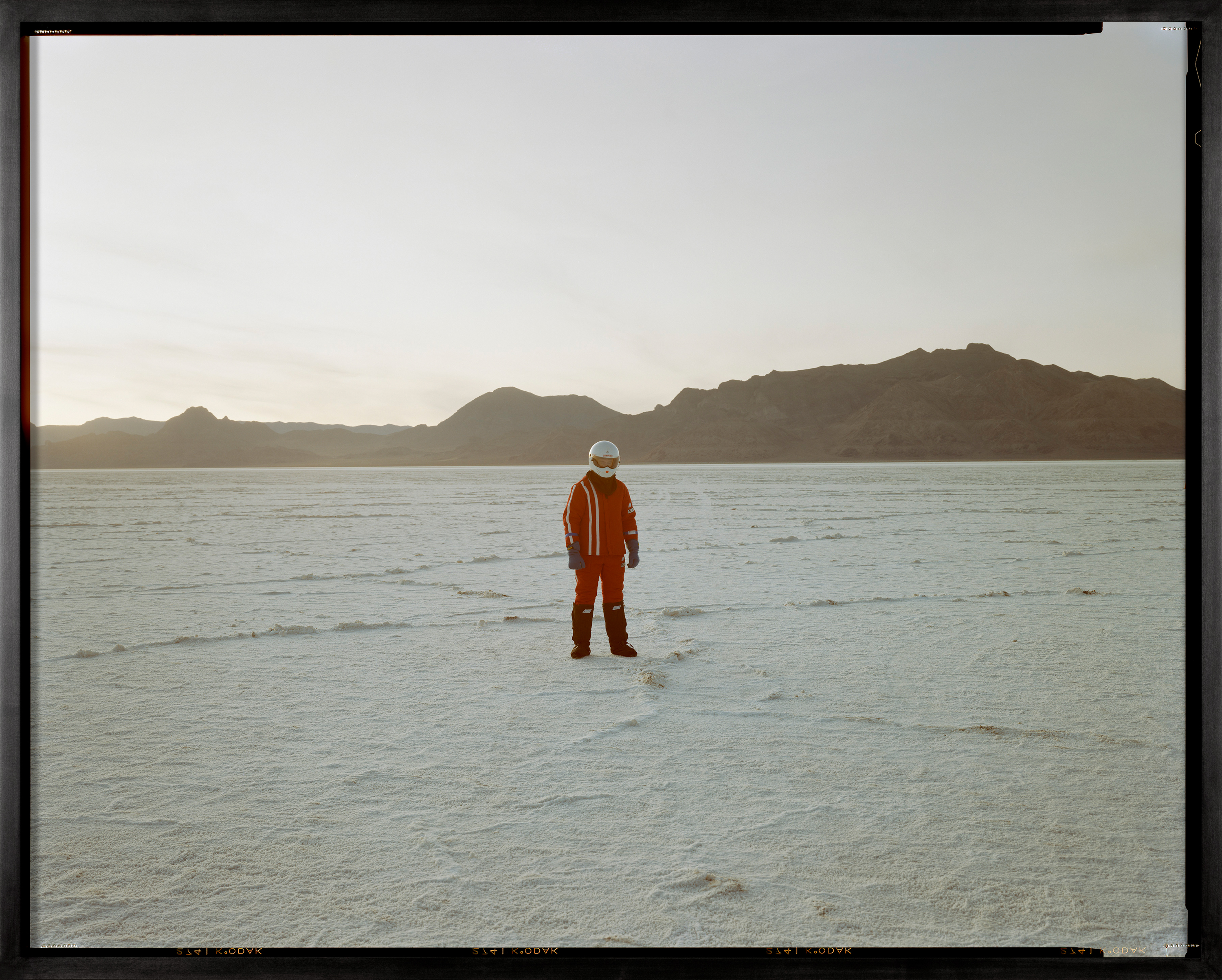 Color image of a color photograph of a figure in a white helmet and red outfit in the middle of salt flats framed in black