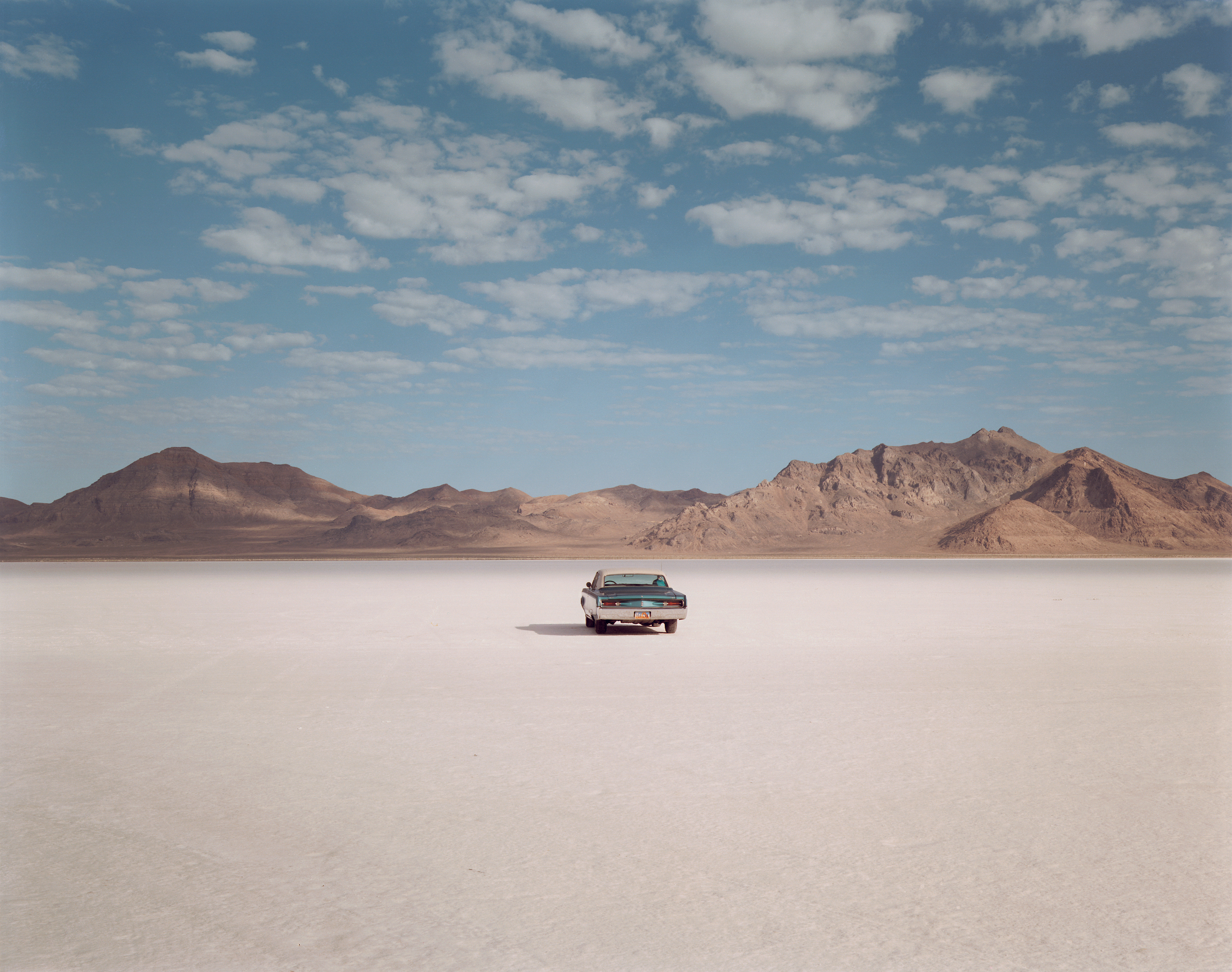Color photograph of a car in the middles of empty salt flats with mountain range along horizon