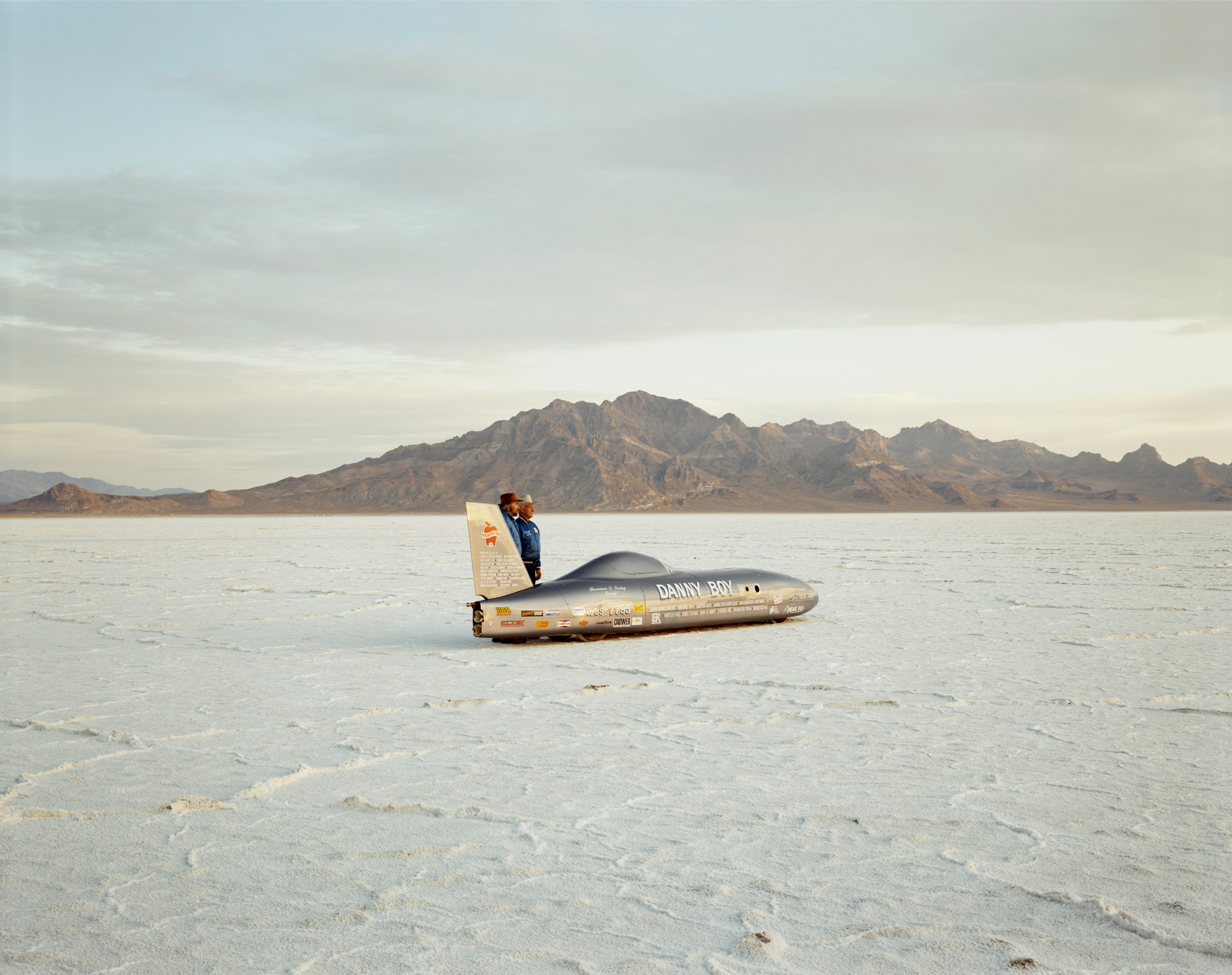 Color photograph of two figures standing by a race car in the middle of salt flats with mountains along the horizon