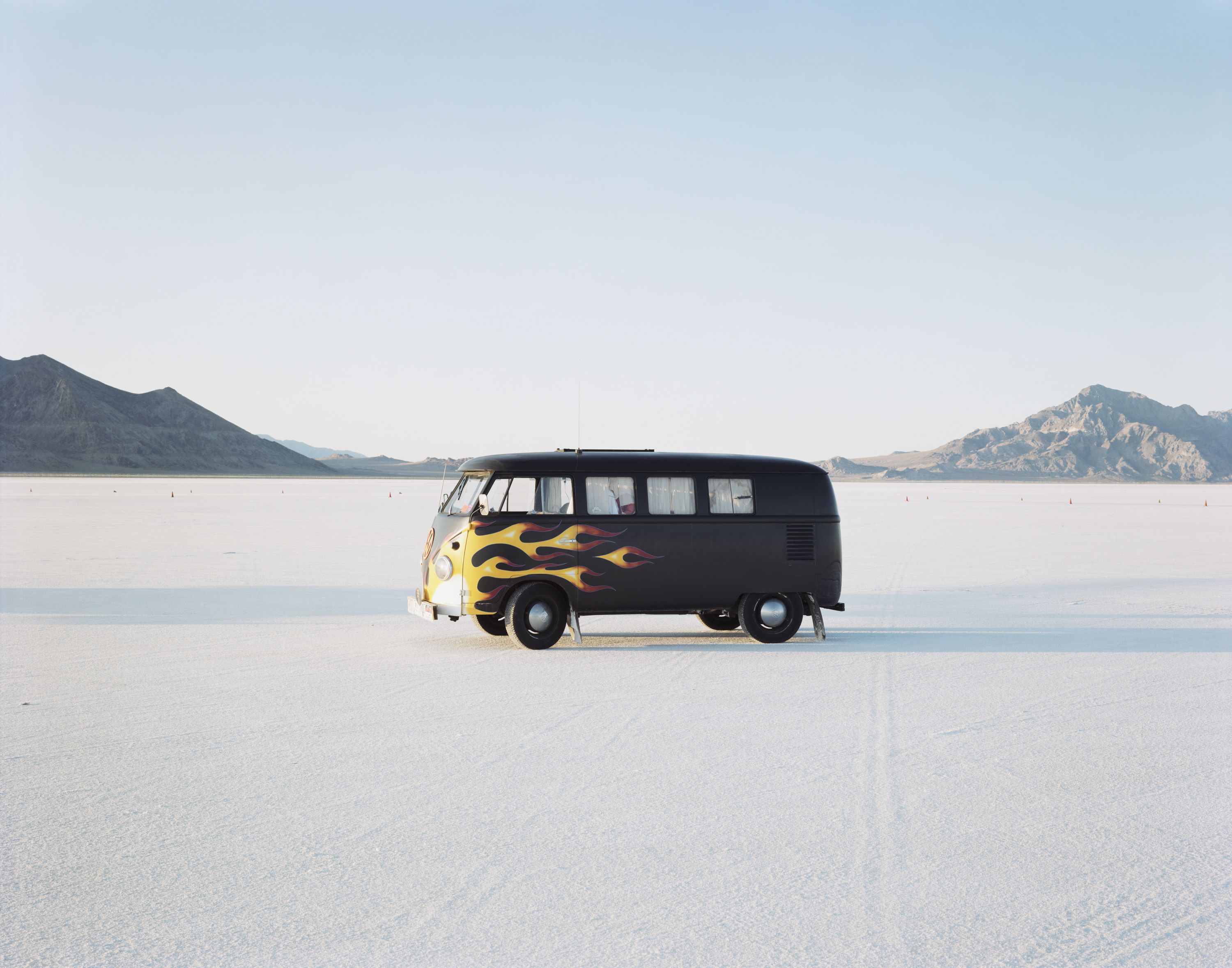 Color photograph of a VW bus with flames painted on the front in the middle of empty slat flats with mountains on horizon