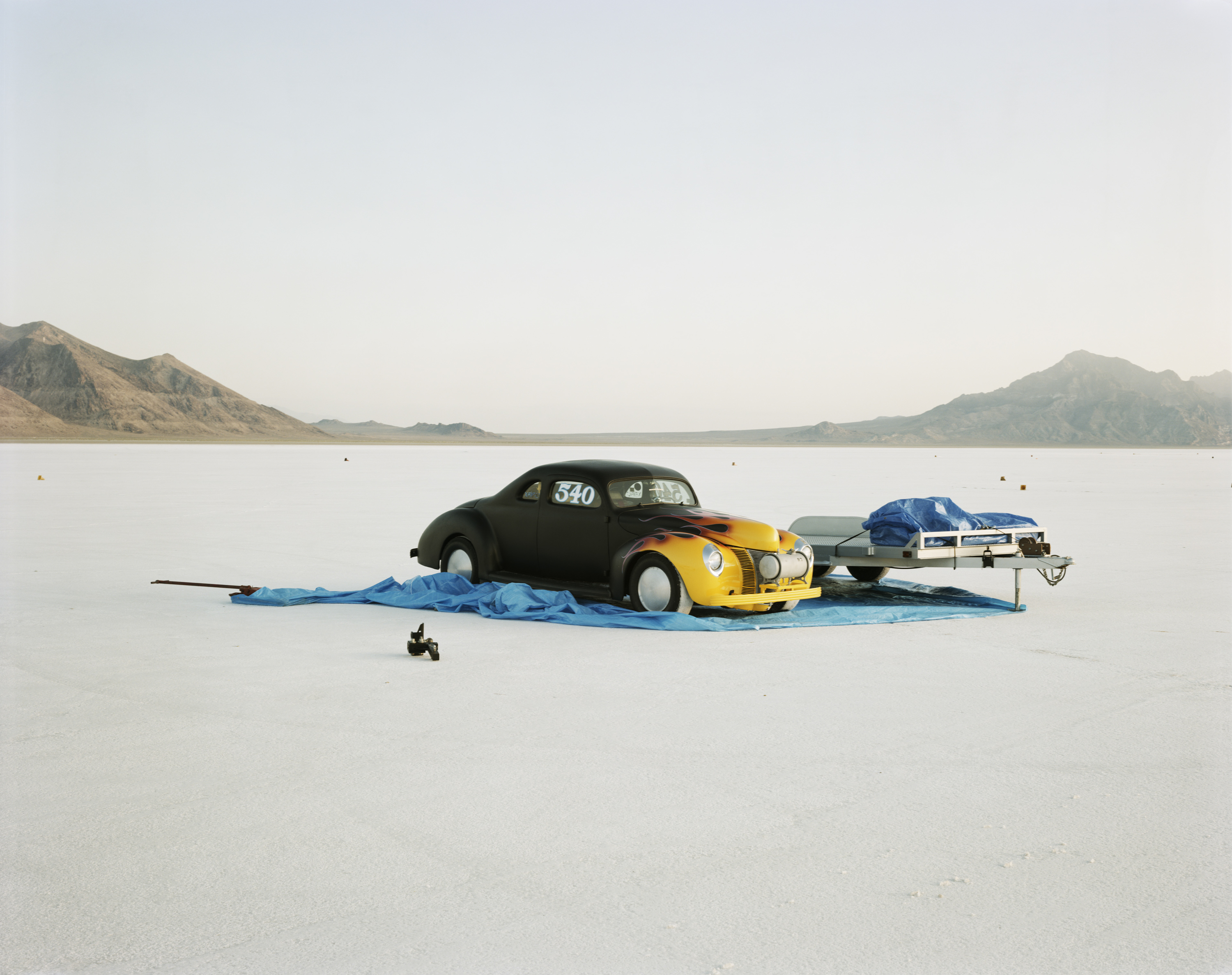 Color photograph of a car with flames painted on the front in the middle of empty salt flats with mountains on horizon