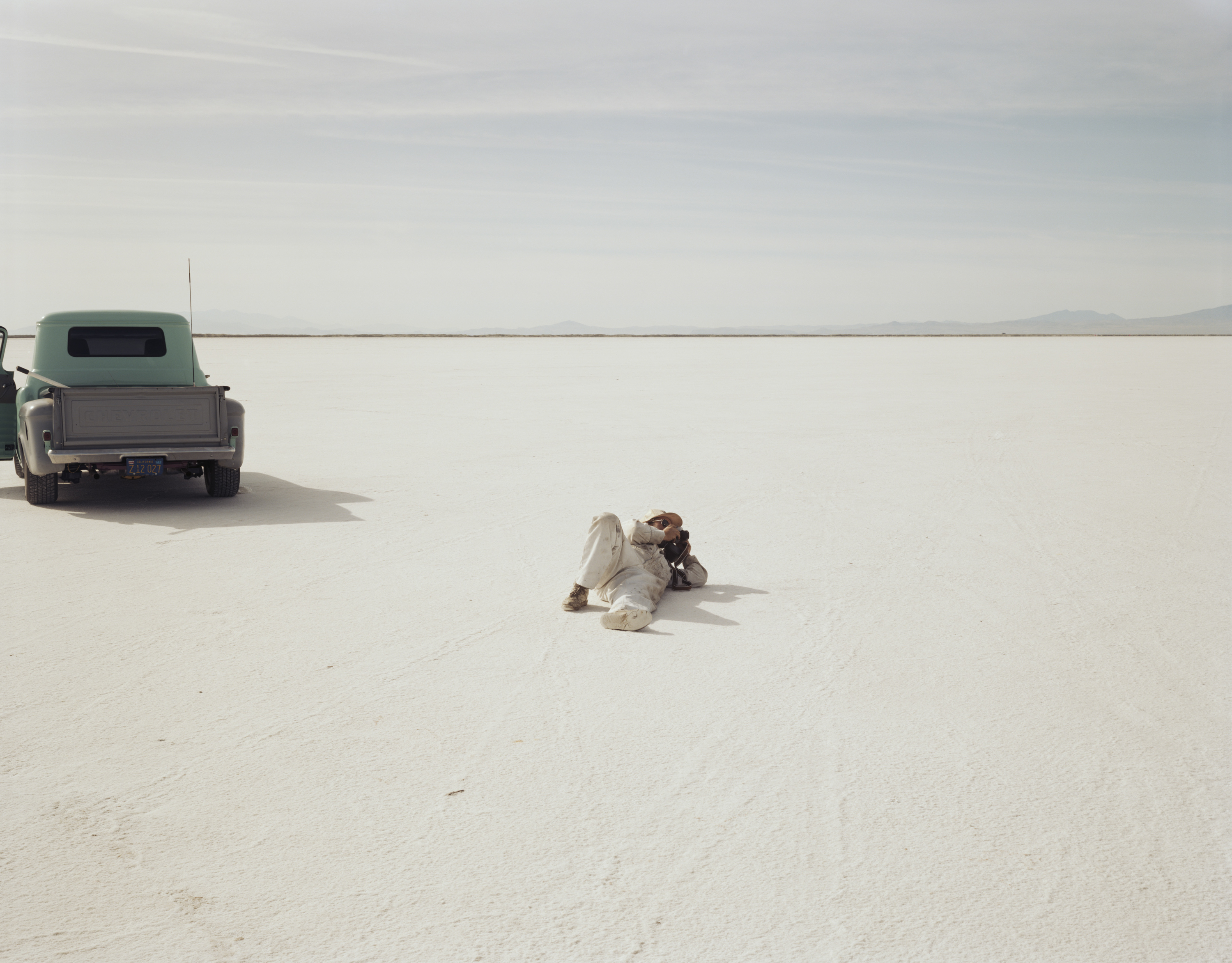 Color photograph of figure laying on the desert ground using a camera near a pickup truck