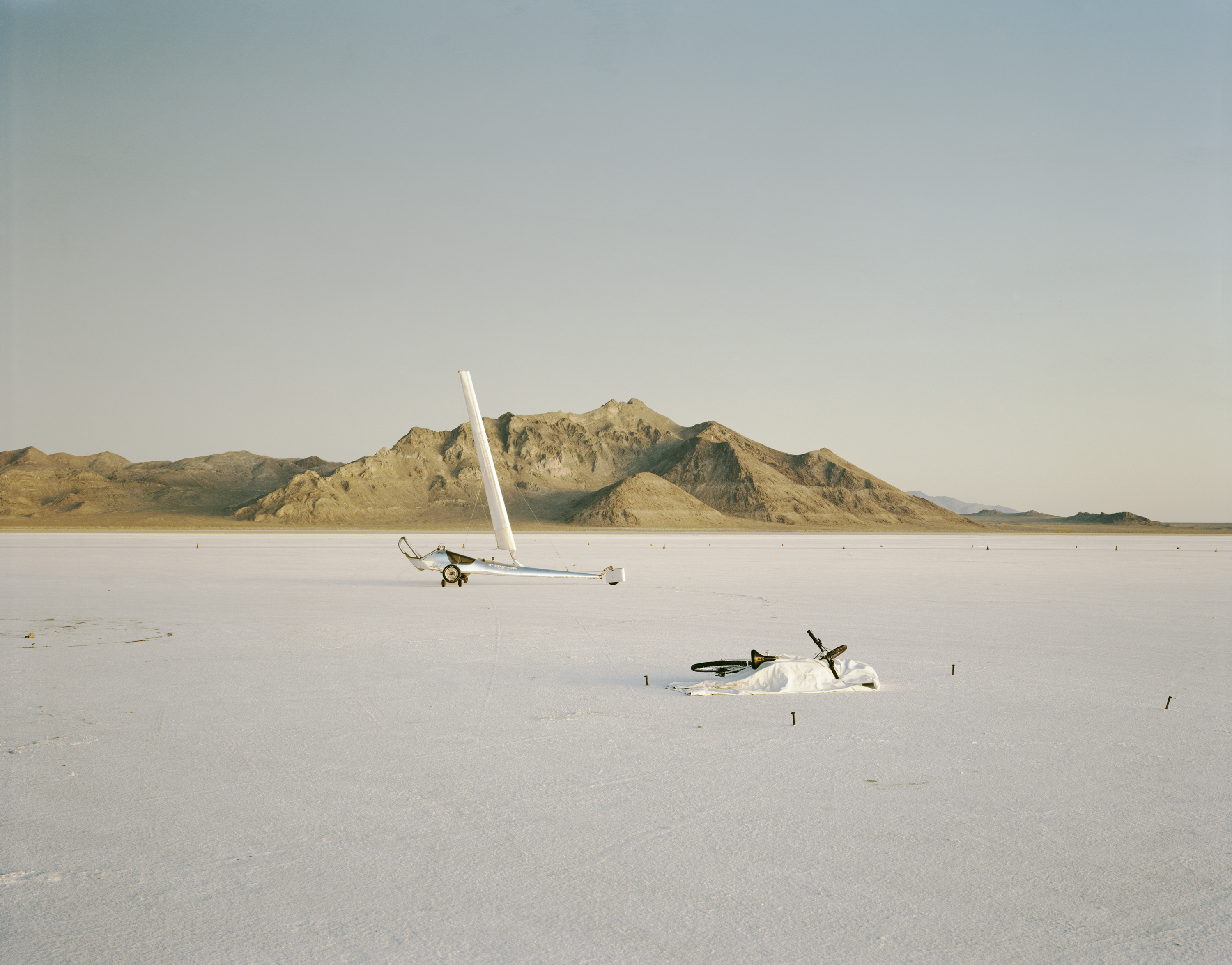 Color photograph of a land sailing vehicle and a bike in the middle of empty salt flats with mountains on the horizon