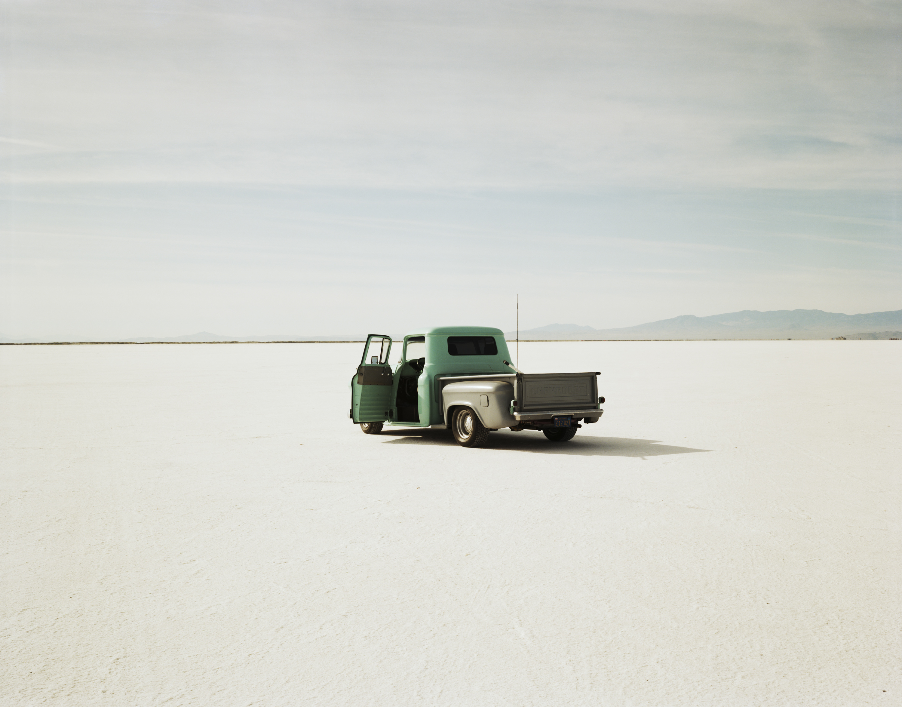 Color photograph of a pickup truck in the middle of empty salt flats with mountains along the horizon