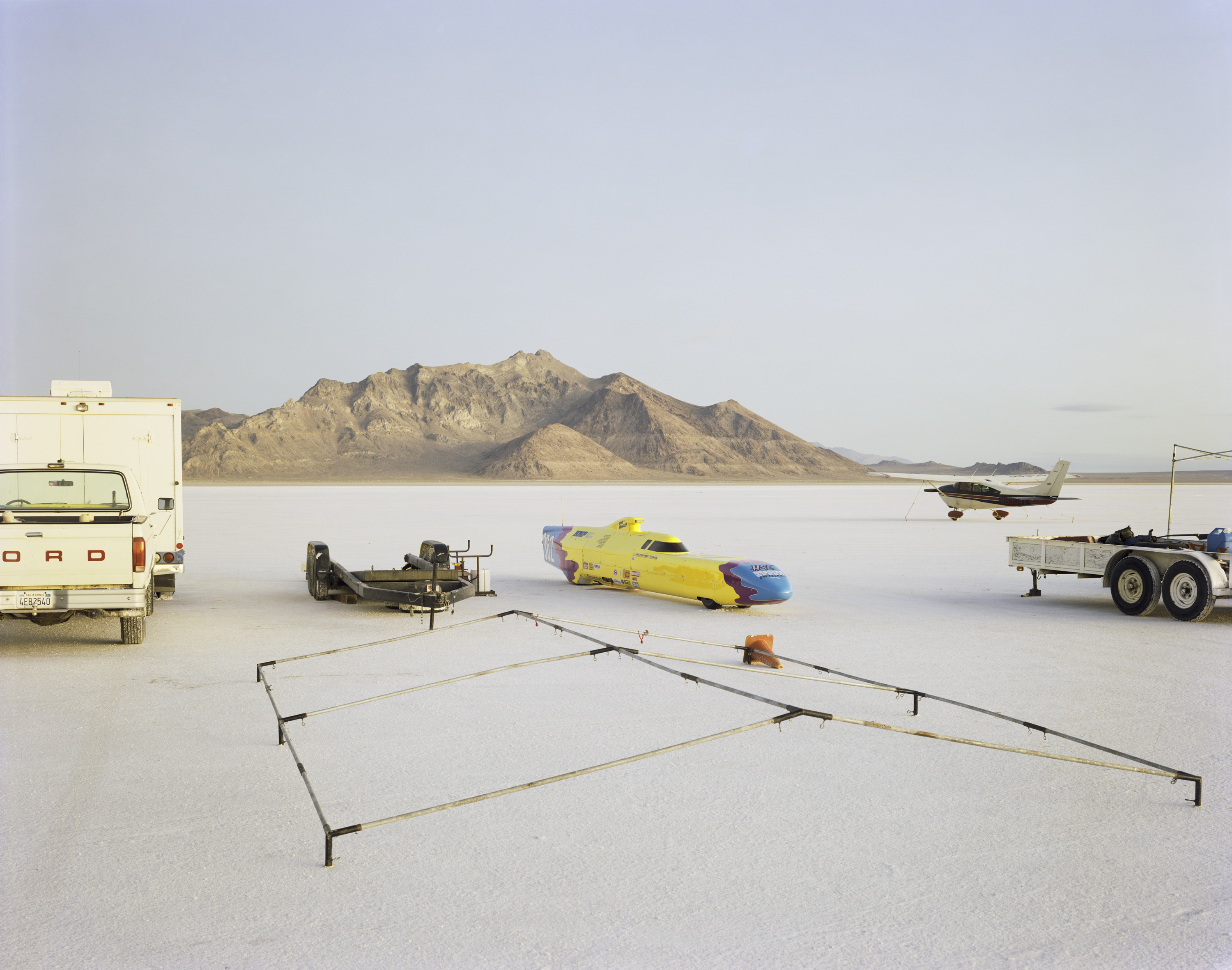 Color photograph of multiple automobiles and a plane in the middle of salt flats with mountains along the horizon