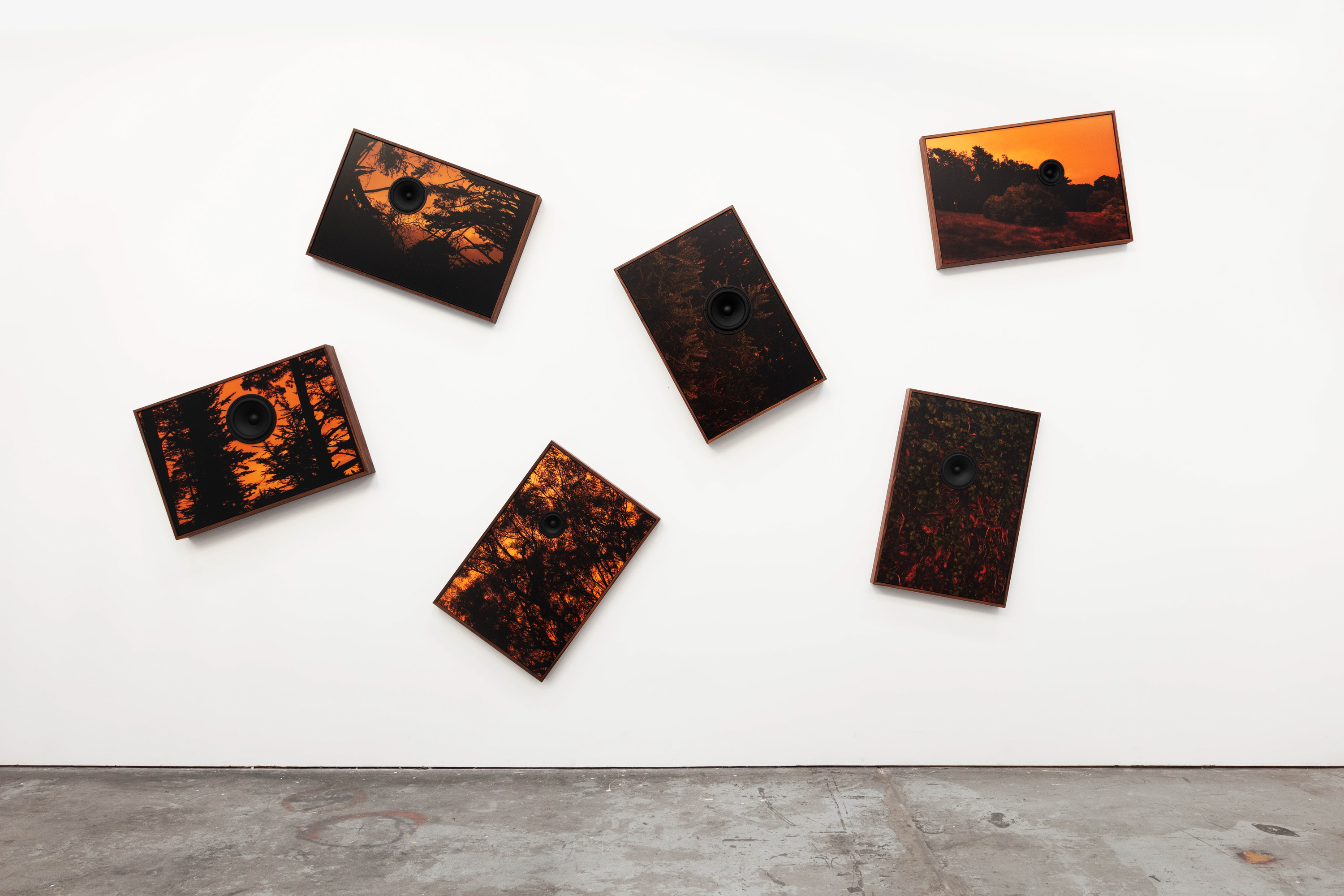 Color image of a suite of six speakers with photographs depicting various orange hued landscapes on white gallery wall