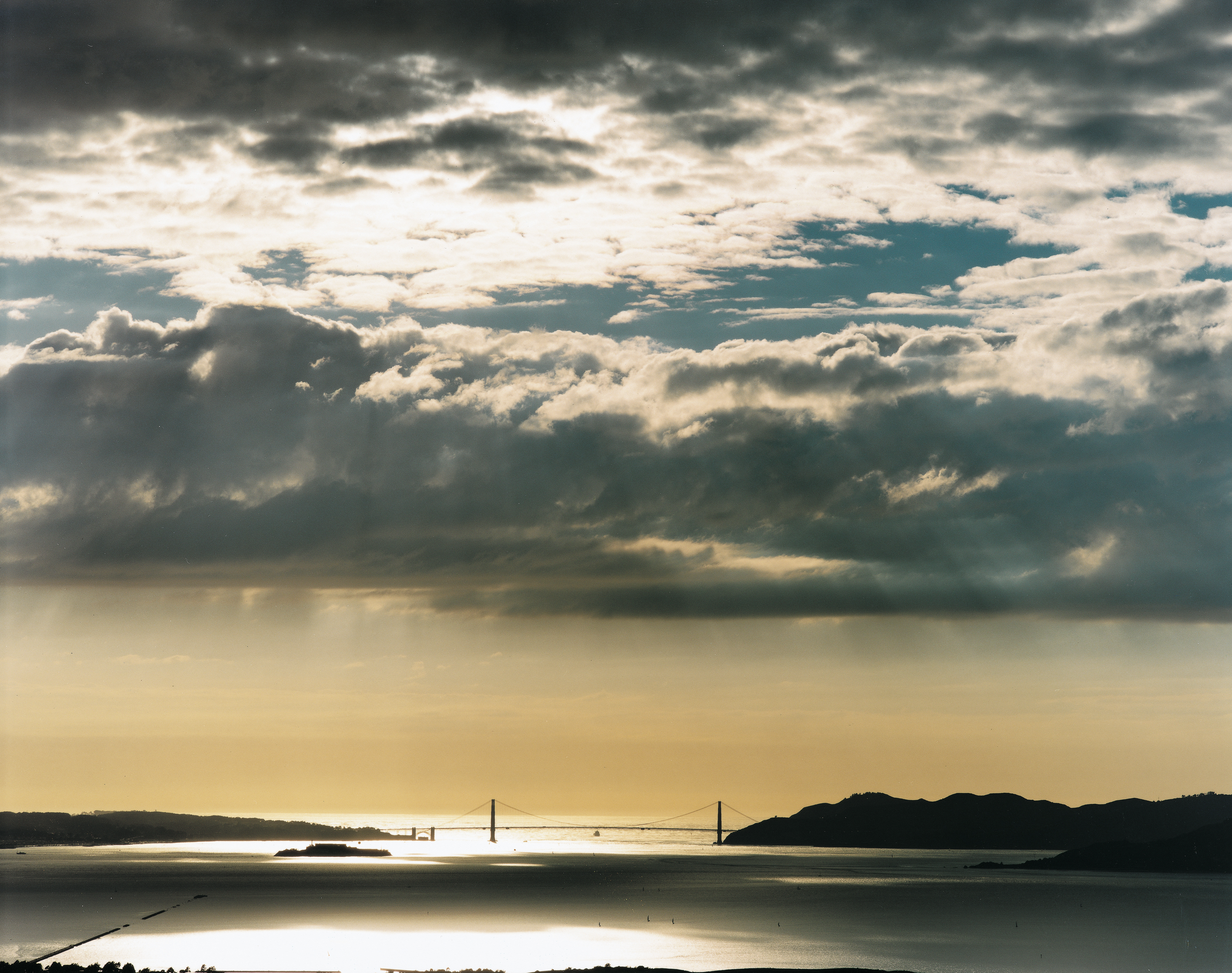 Color photograph of San Francisco Bay and Golden Gate Bridge with sun flooding through a cloudy sky