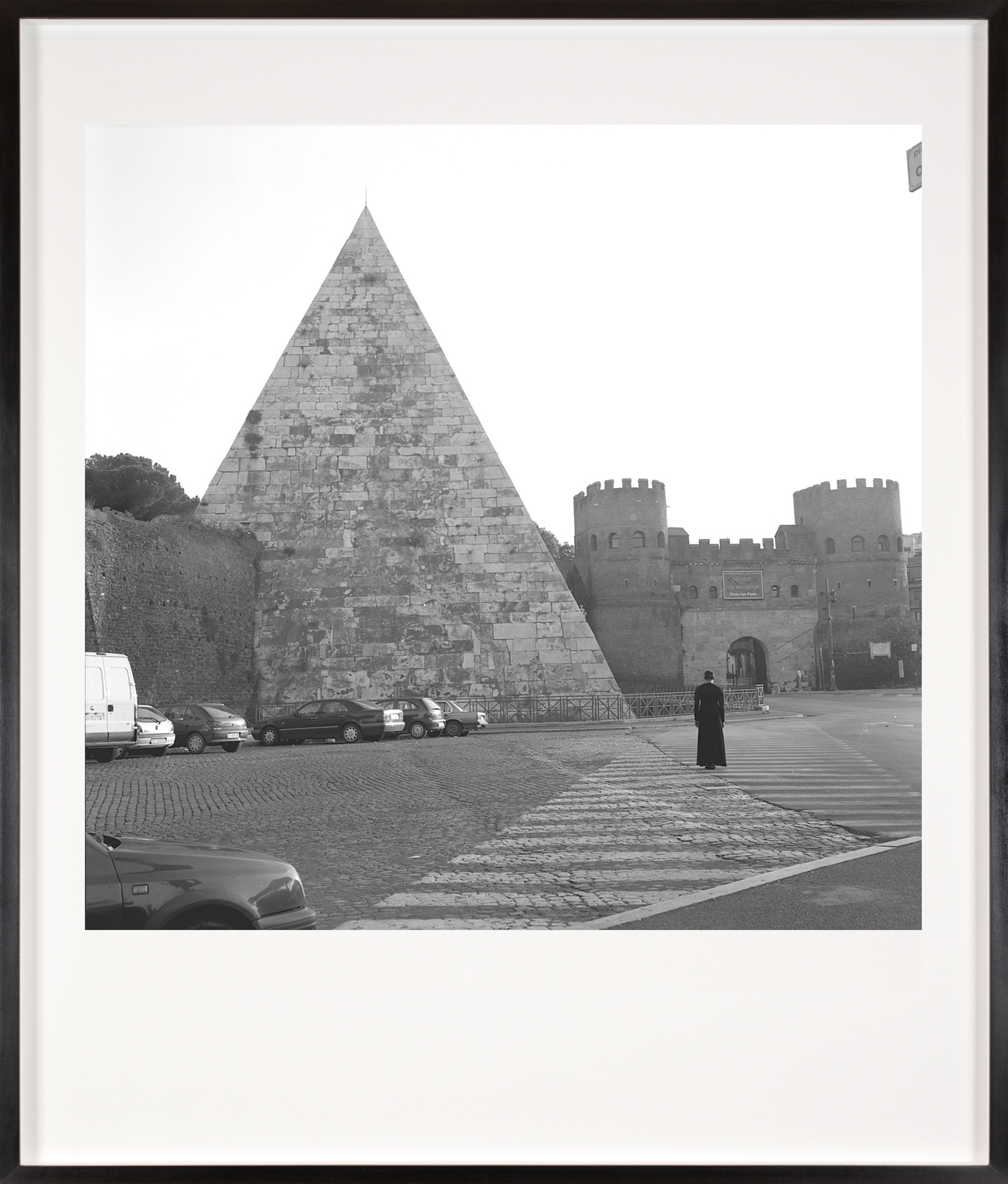 Black and white photograph of a figure in black looking at a pyramid within a city from the crosswalk framed in black
