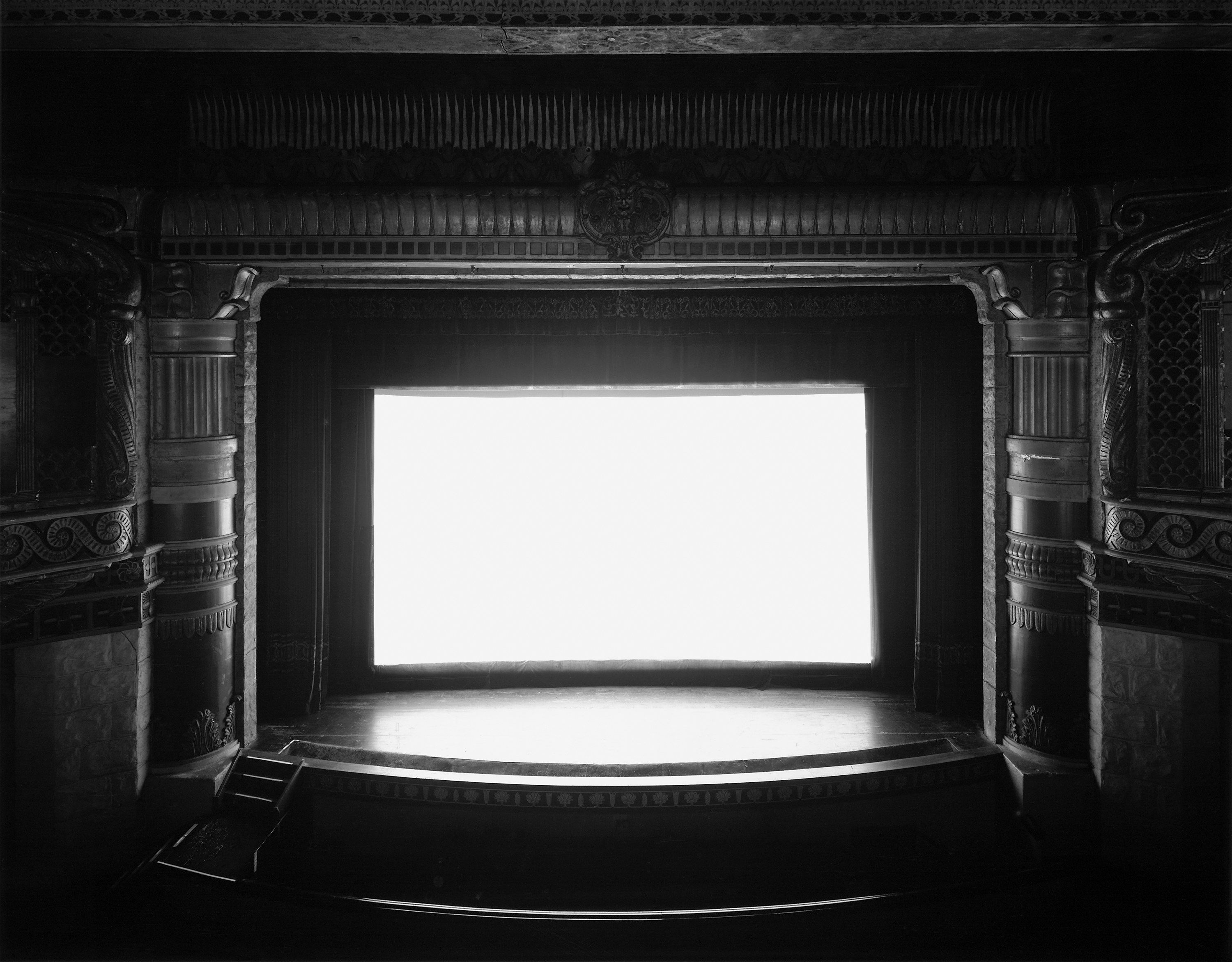 Black and white photograph of an empty theater with a blank white screen illuminating the space