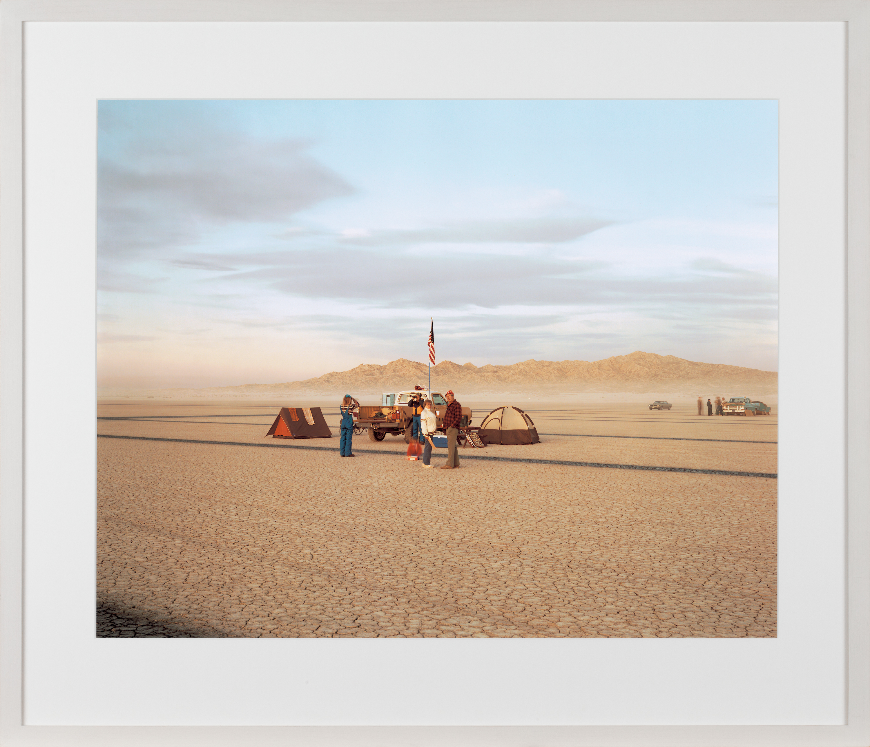 Color image of a color photograph depicting figures in the middle of a desert near tents and cars framed in white