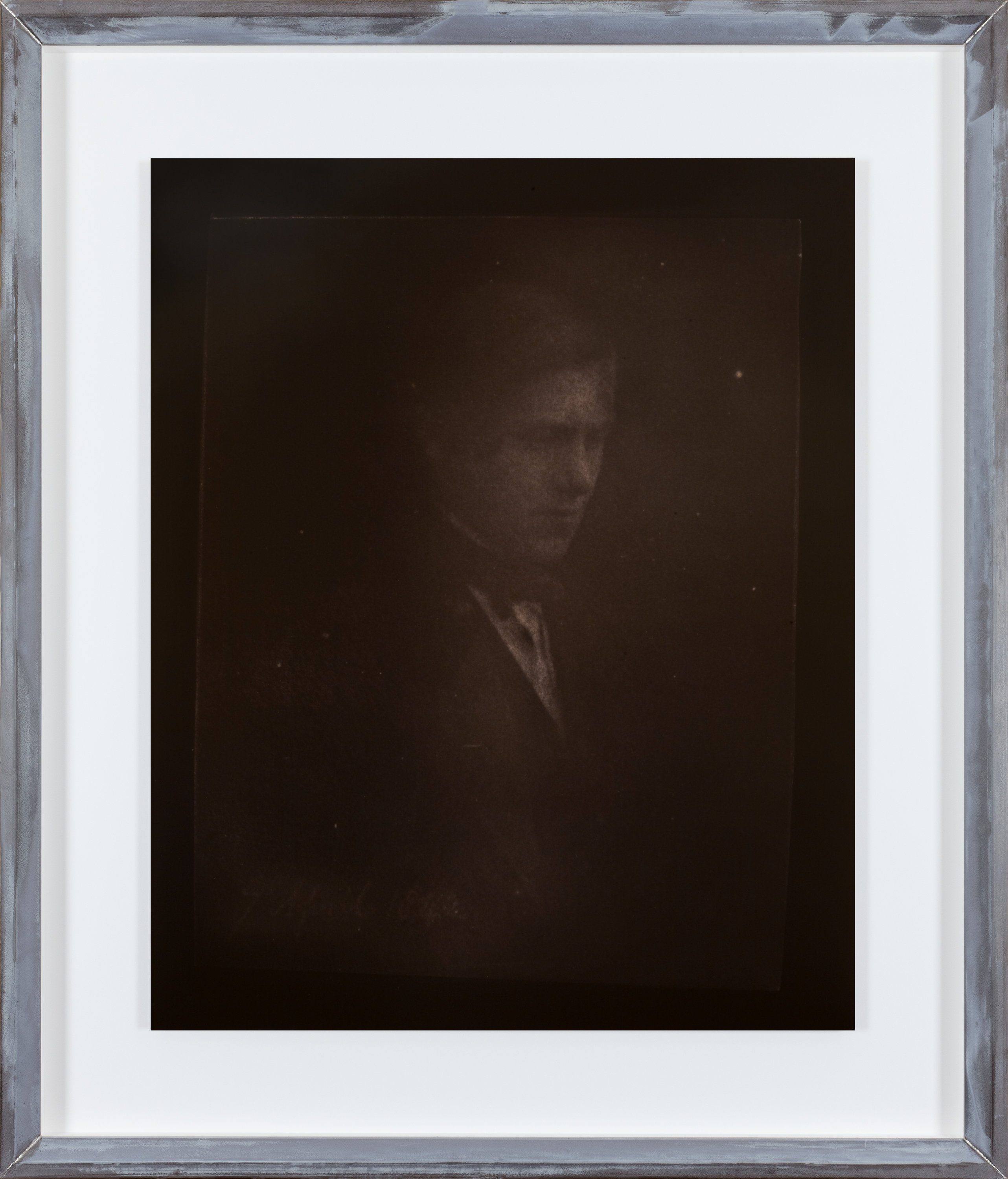 Color photograph of sepia toned portrait of a formal dressed figure with a black background in an aluminum frame