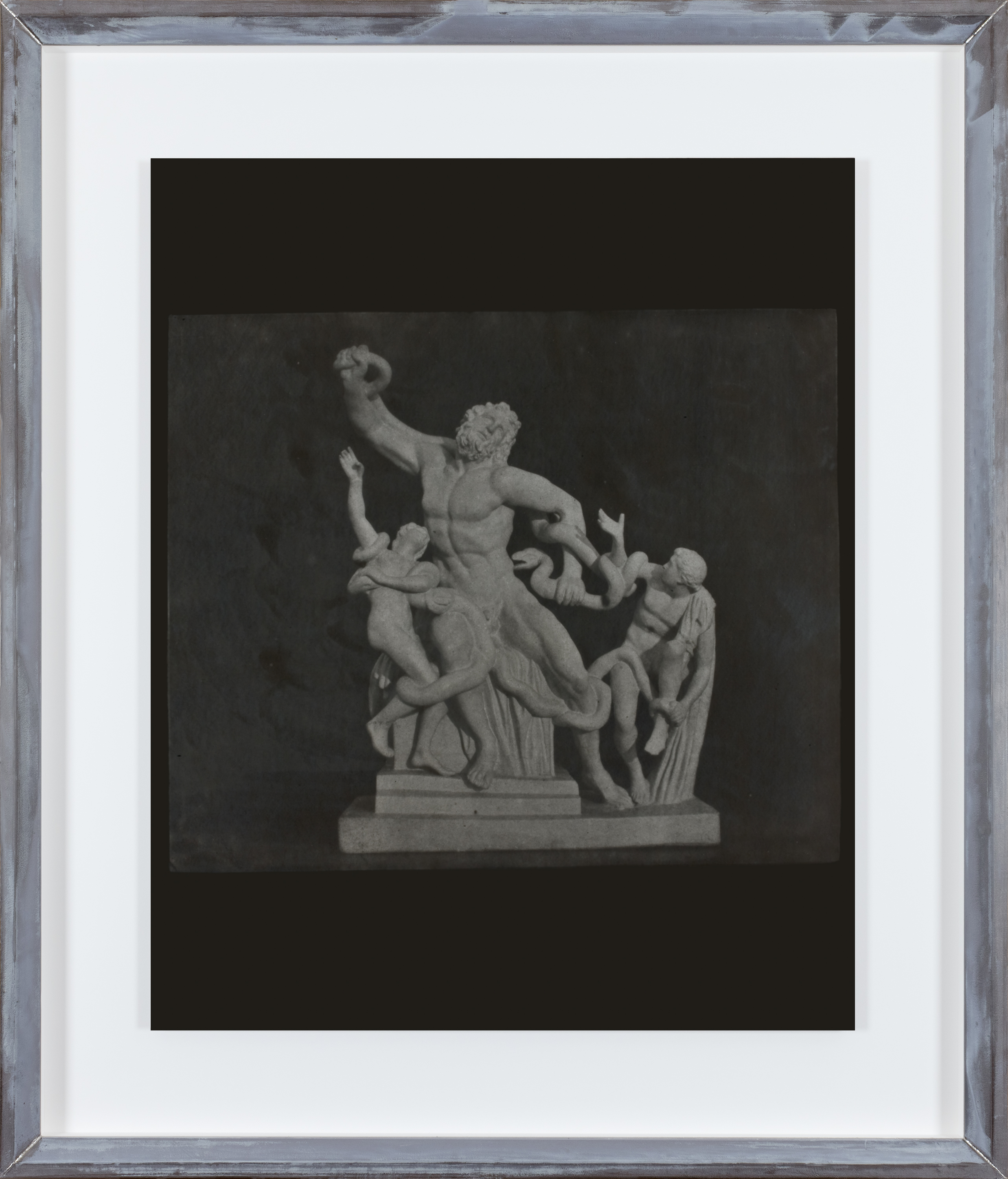 Color photograph of a grey toned sculpture with three figures wrapped around a snake with a black background in an aluminum frame