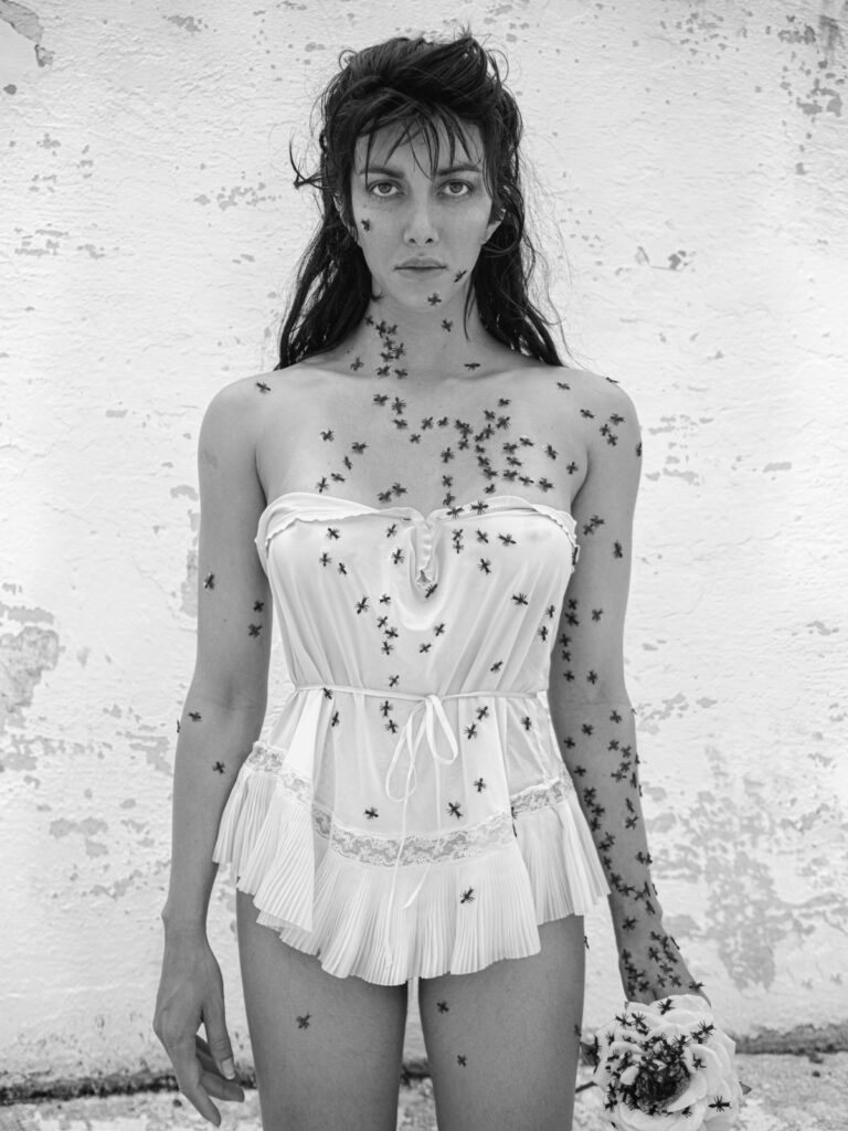 A black and white photograph shows a woman in a white negligee against a wall with chipped white paint. Small insects crawl on her body and on the flower-like object she holds in her hand.