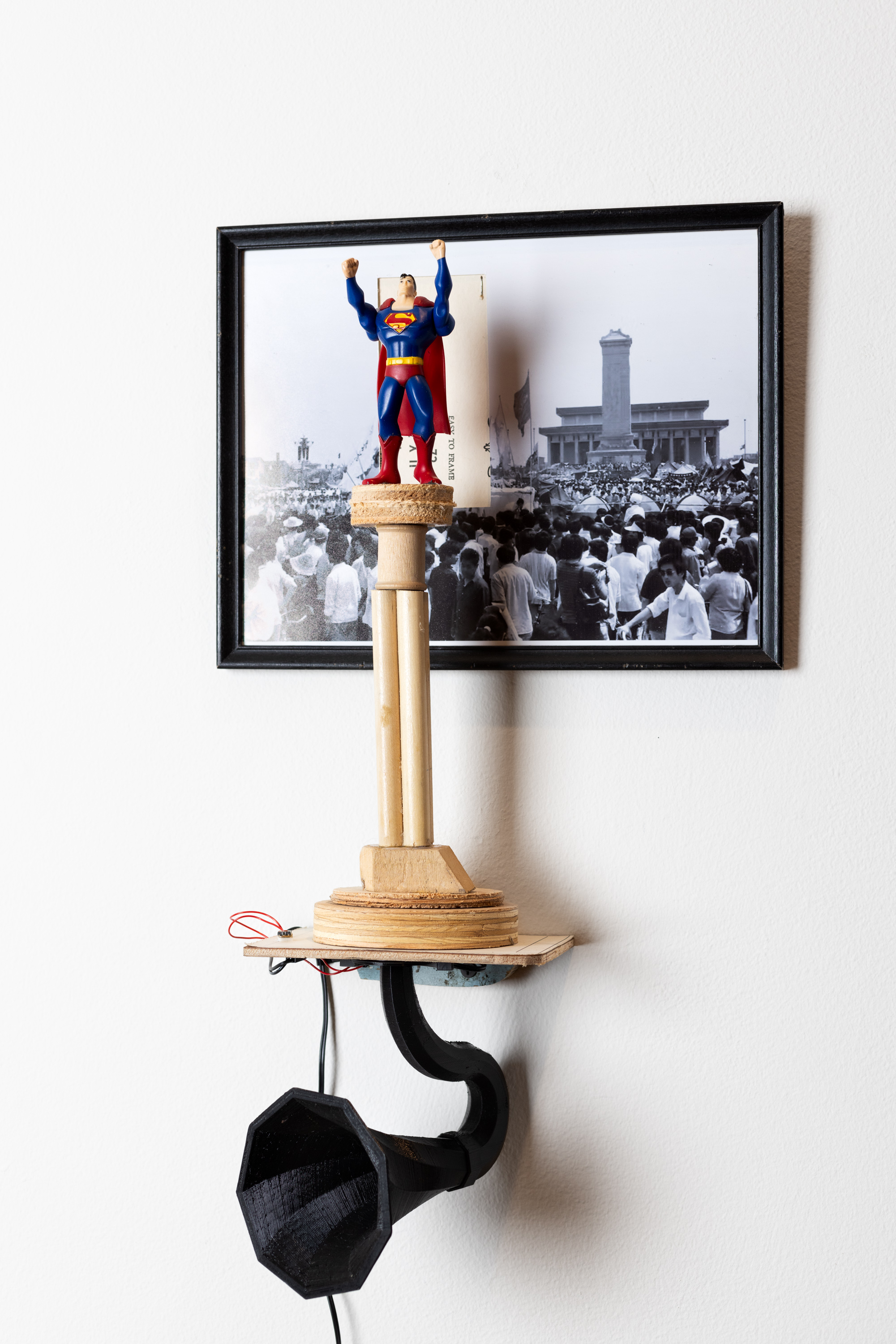 Color image of a mixed media artwork with a framed black and white photograph along with a small plastic superman figure on a wooden pedestal with a small gramophone