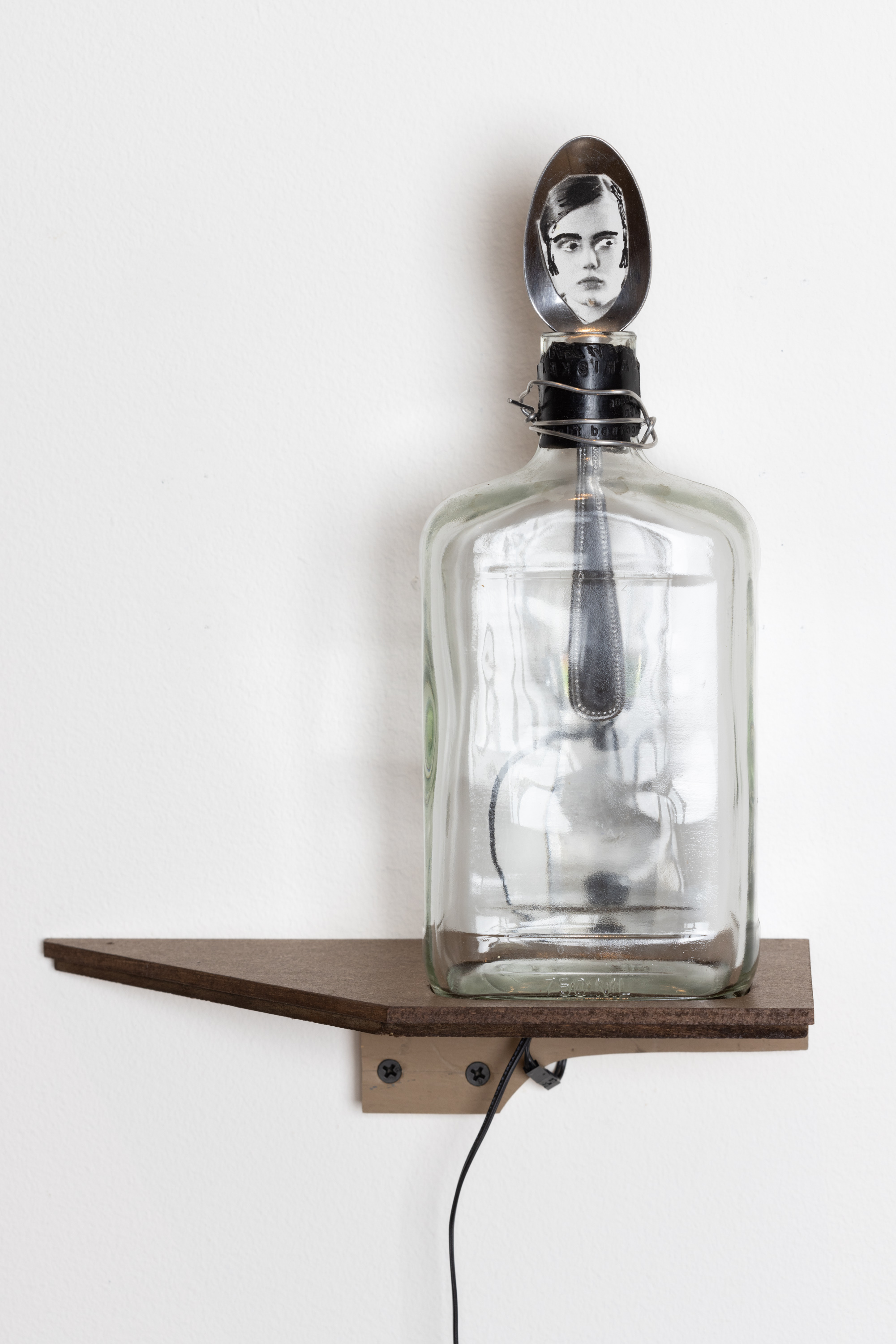 Color image of a mixed media artwork consisting of a glass bottle on a wooden shelf with a silver spoon inserted in the bottle