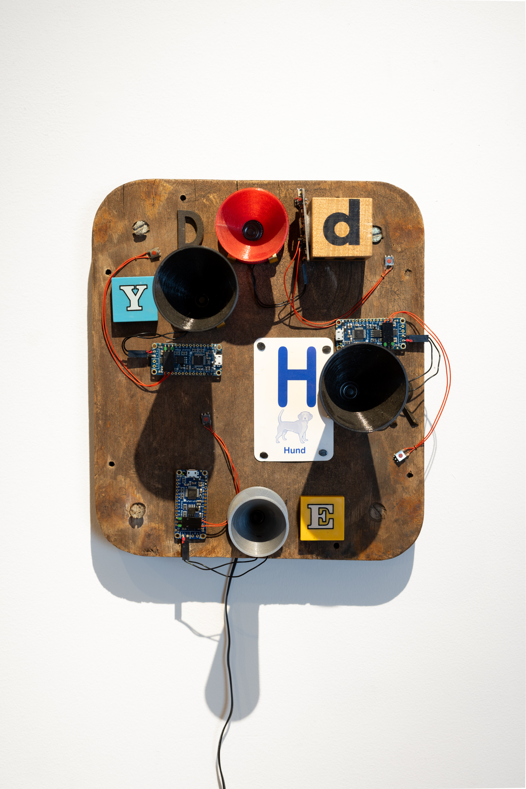 Color image of a mixed media artwork consisting of four speakers and exposed wiring on an aged piece of wood with found objects attached to the face of the work