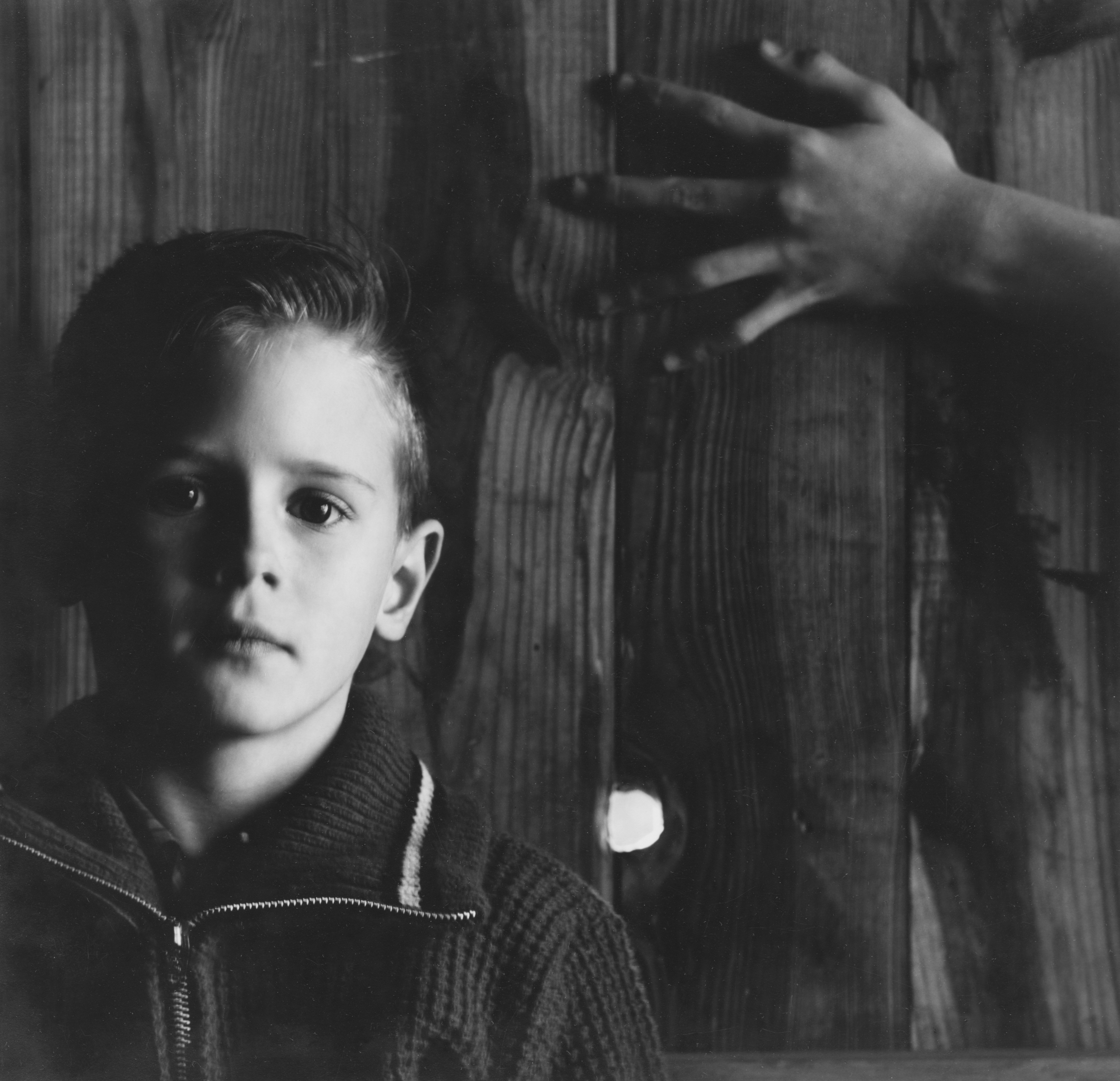 Black and white photograph of a child in front of wooden wall with a hand extended on top right of frame