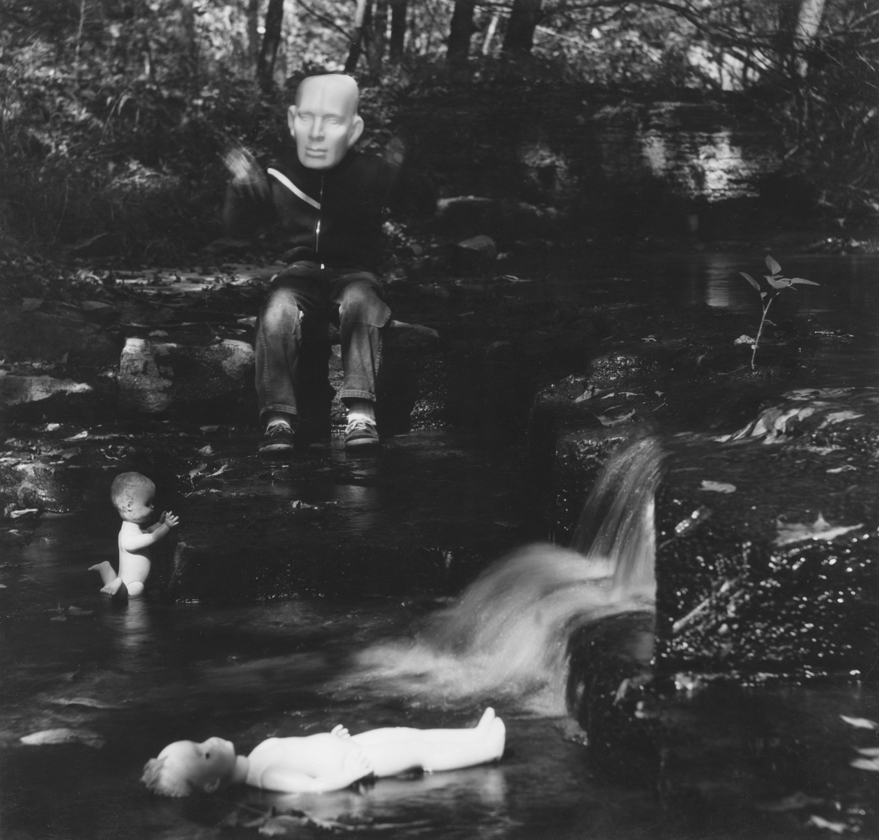 Black and white photograph of a child figure wearing a mask and sitting by a creek with dolls in the water