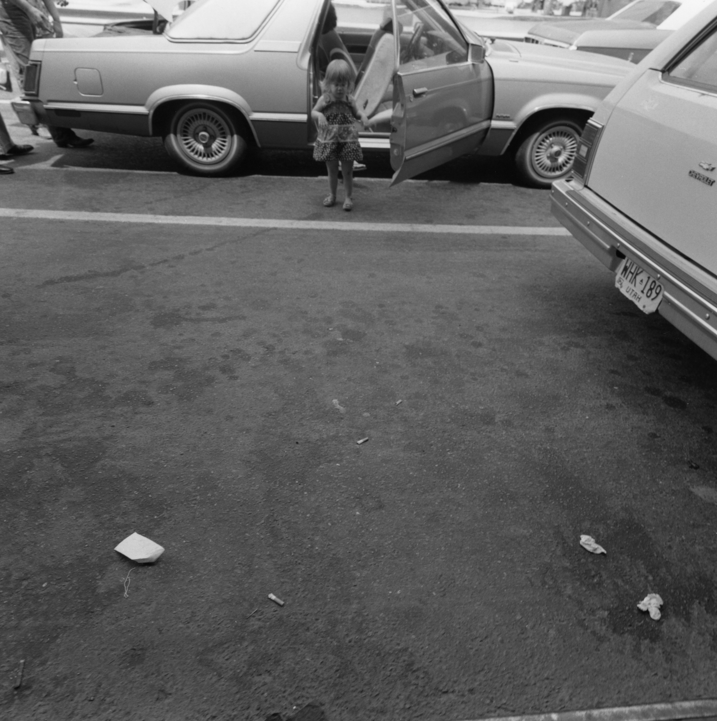 Black and white photograph of a child exiting a car and standing in a parking lot with littered debris