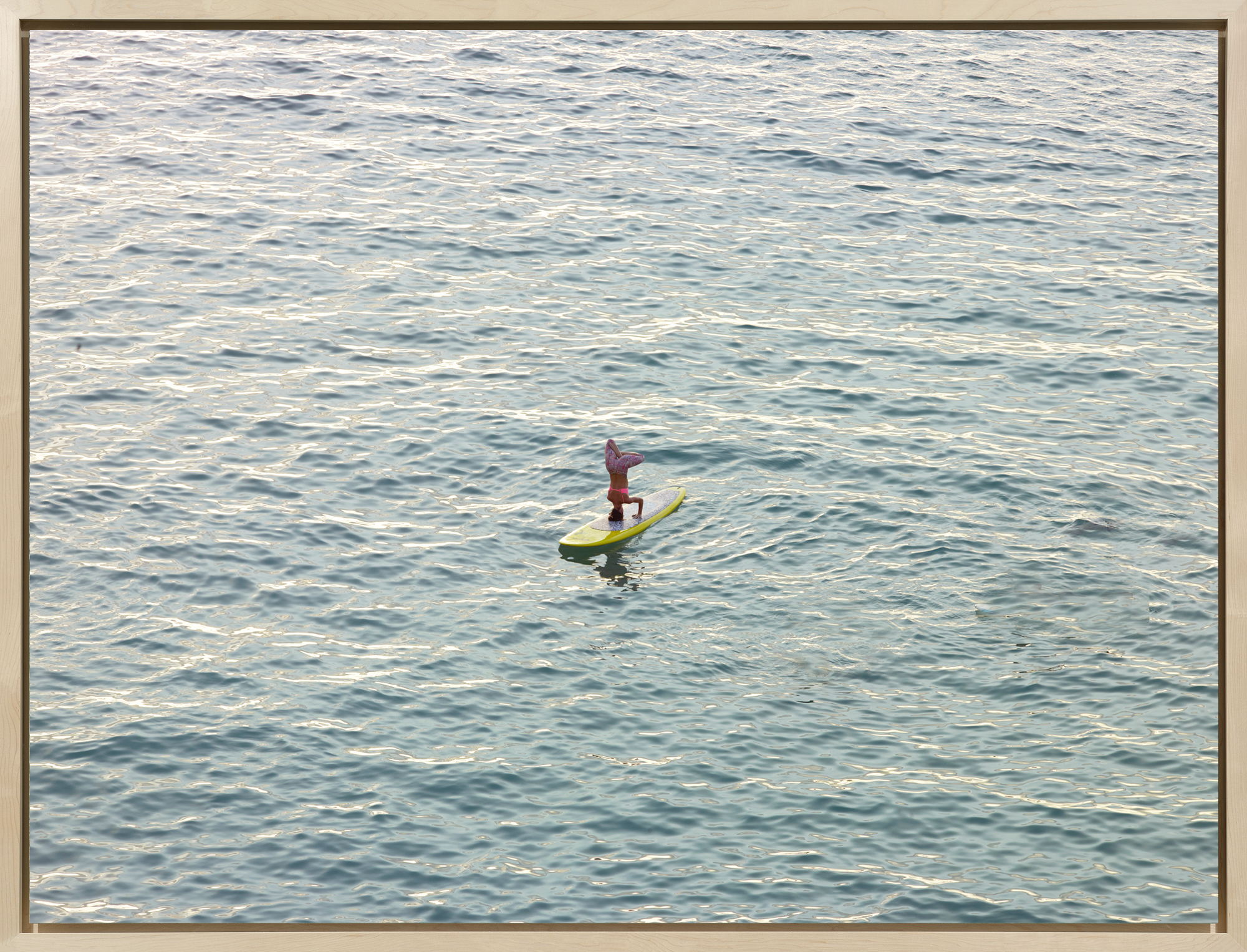 Color photograph of a woman doing a headstand atop a surfboard, afloat on a calm ocean surface.