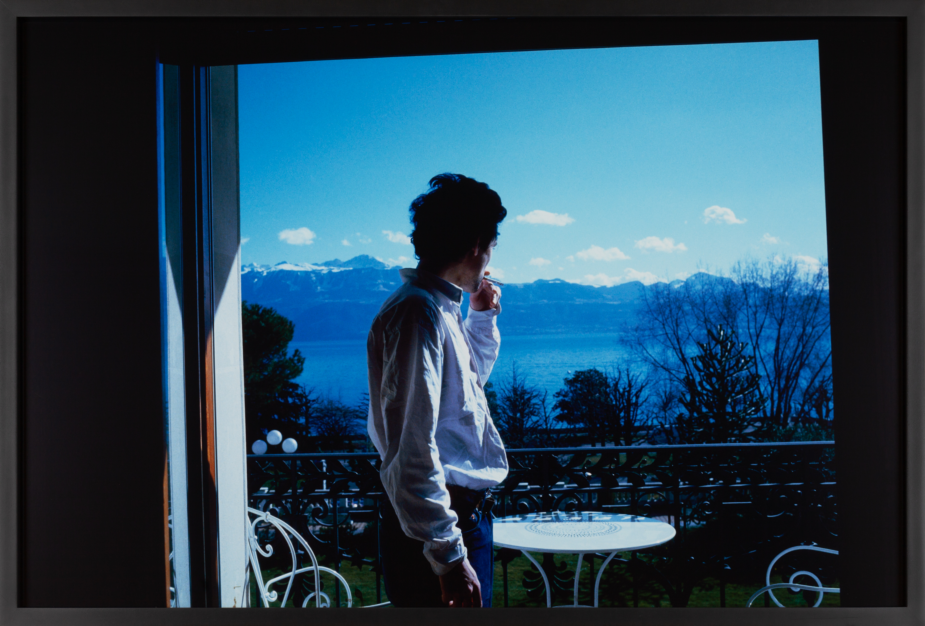 Color photograph of a figure smoking on a balcony and looking towards the horizon framed in black