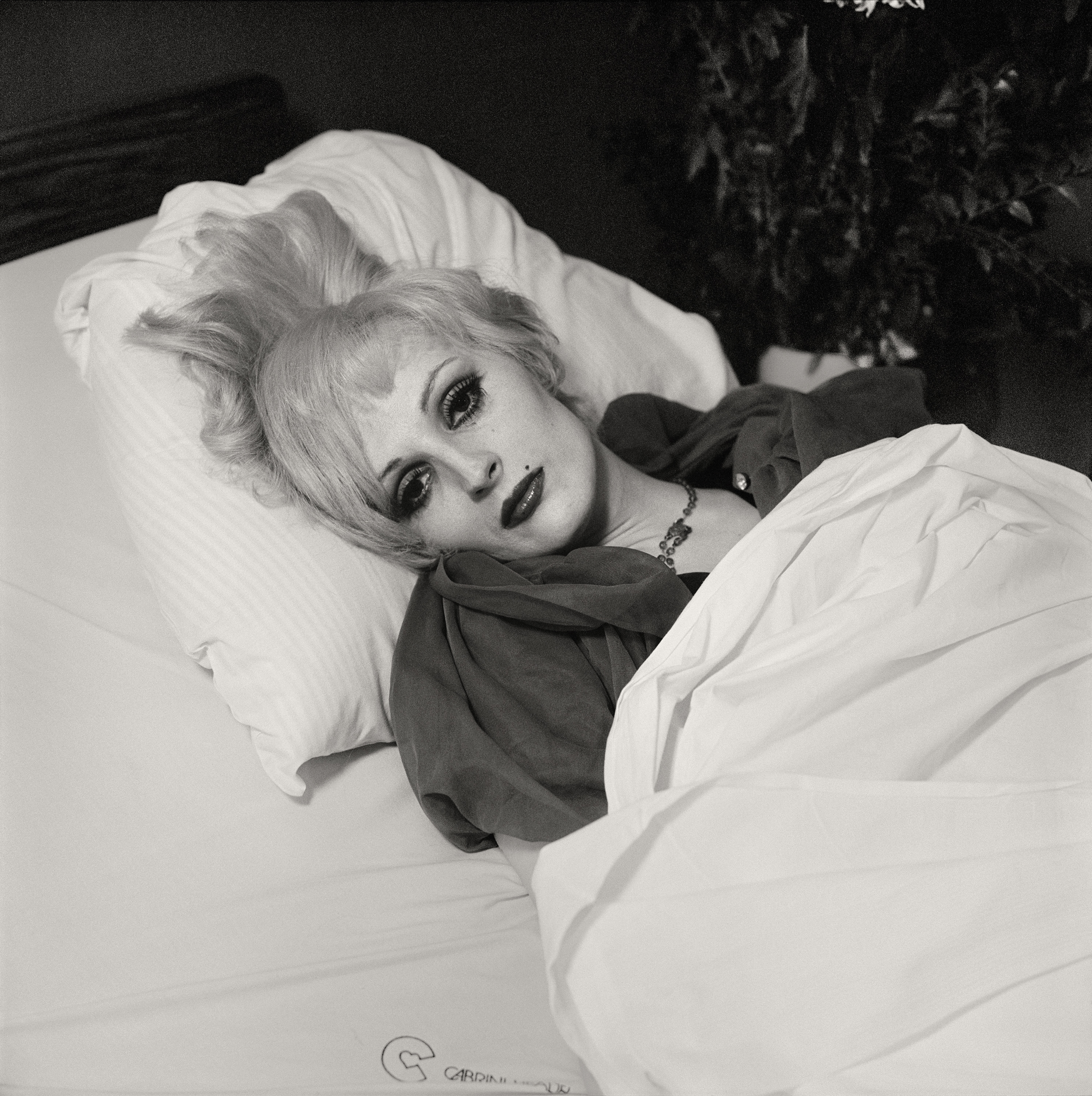 Black and white photograph of a female figure in a hospital bed wearing a full face of makeup and arms at their sides underneath white sheet