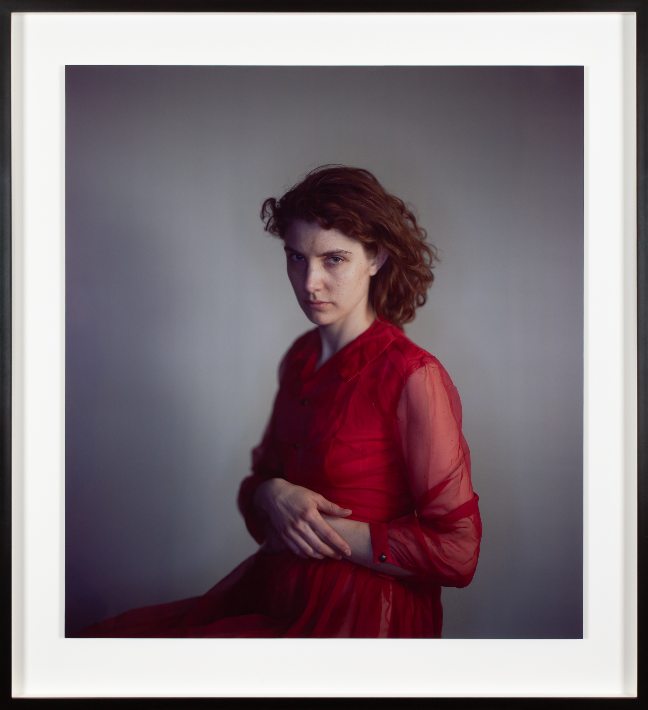 Color photograph of a seated figure in red dress gazing at the viewer framed in black