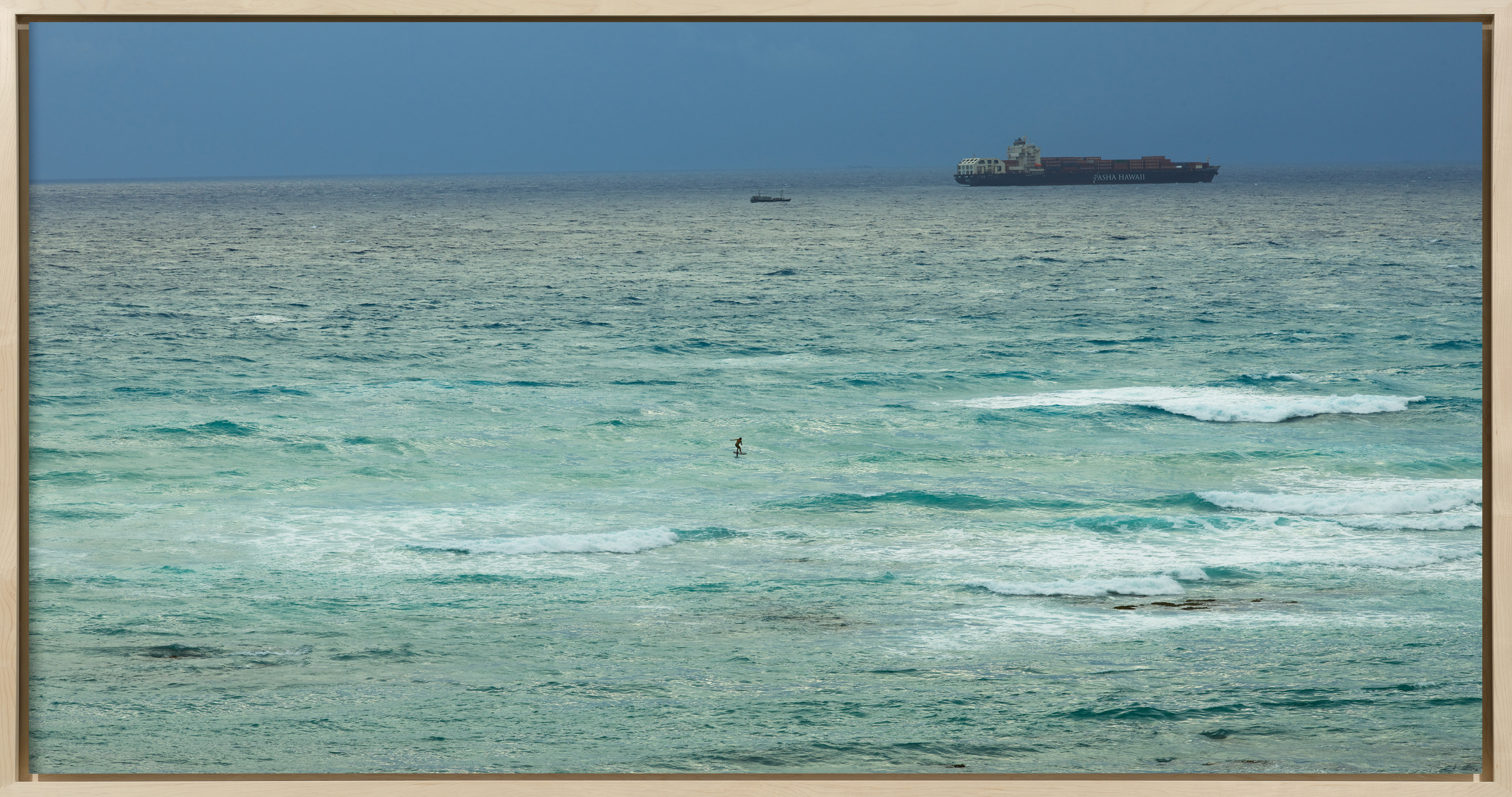 Color photograph of a figure on a hydrofoil surfboard in the middle of choppy blue waters with cargo ship on the horizon framed in bleached wood
