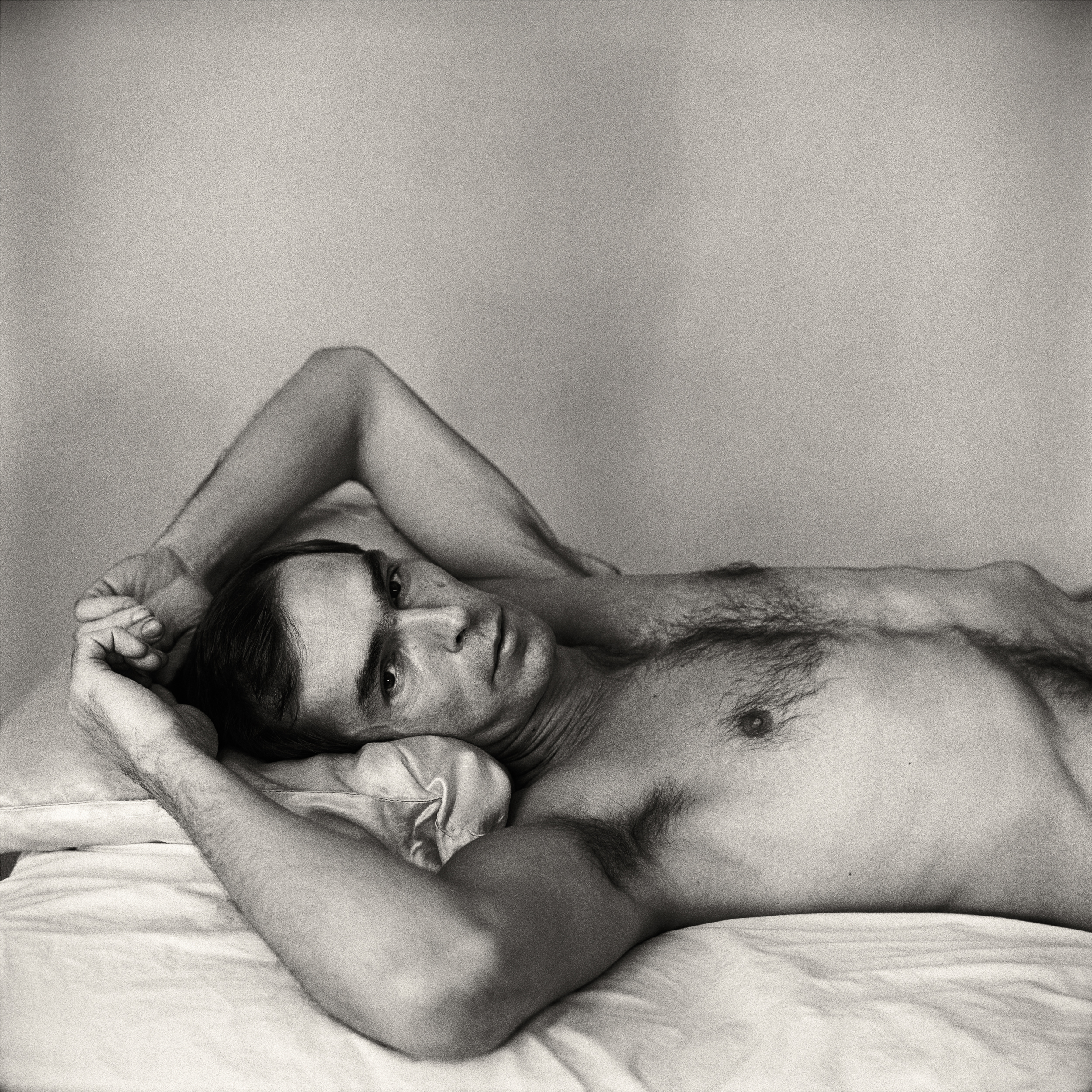 Black and white photograph of a shirtless dark-haired man reclining with his hands in his hair