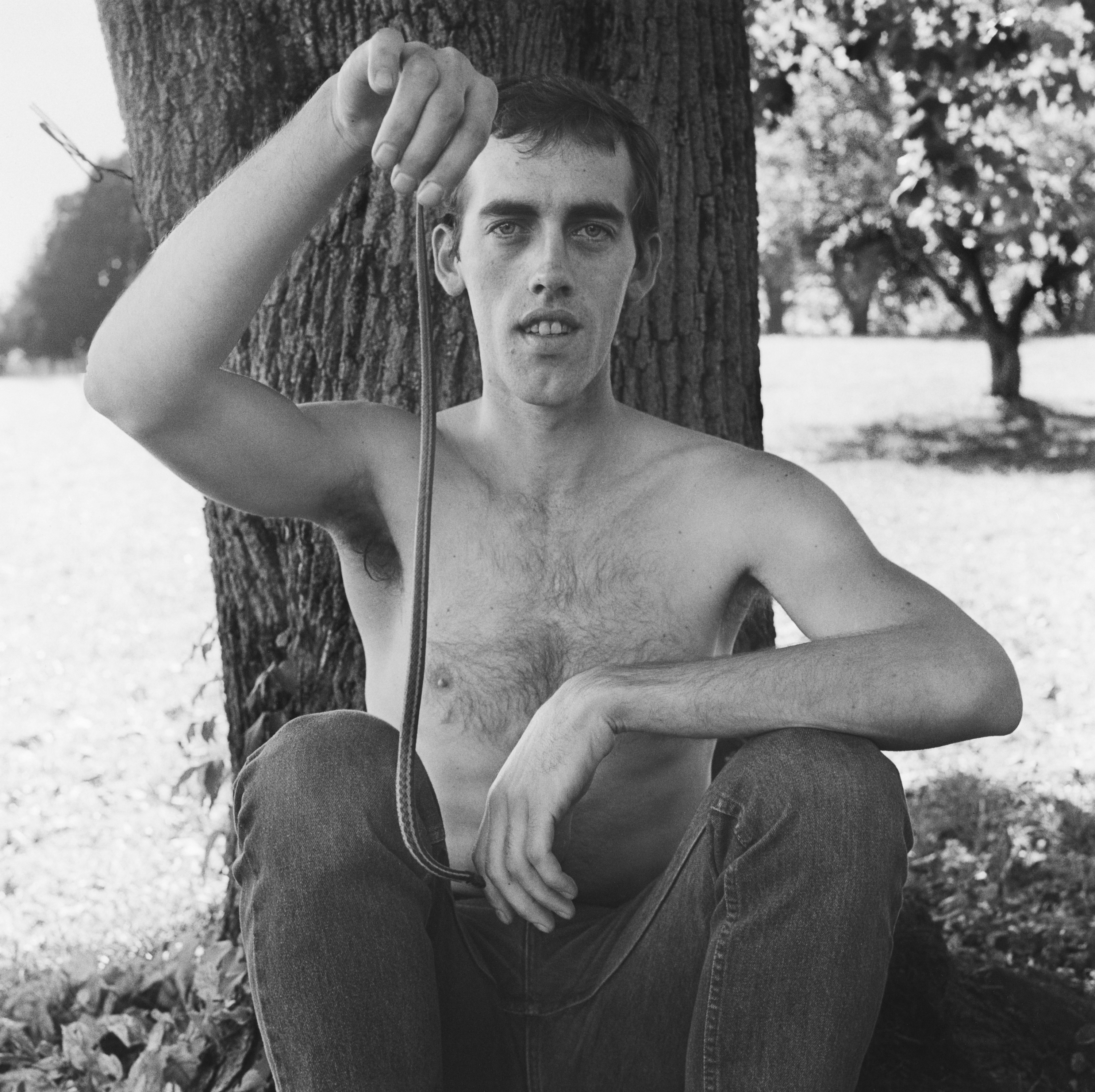 Black and white photograph of a shirtless male figure seating at the base of a tree and holding a snake
