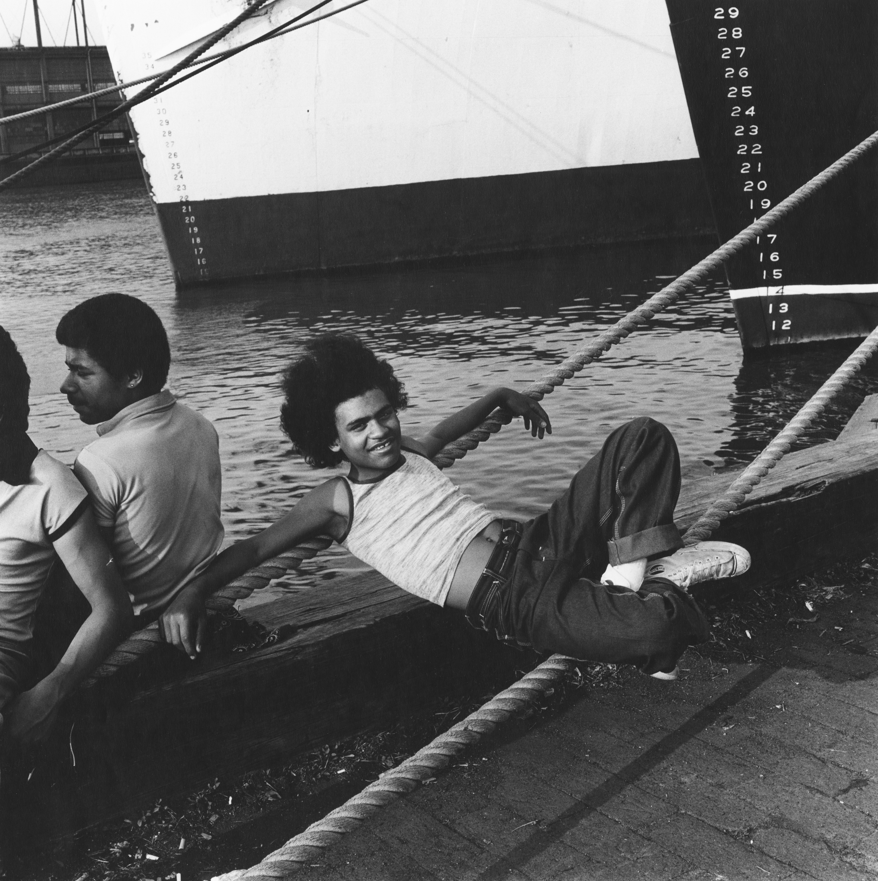 Black and white photograph of a figure laying on ropes attached to a pier while looking directly at the viewer