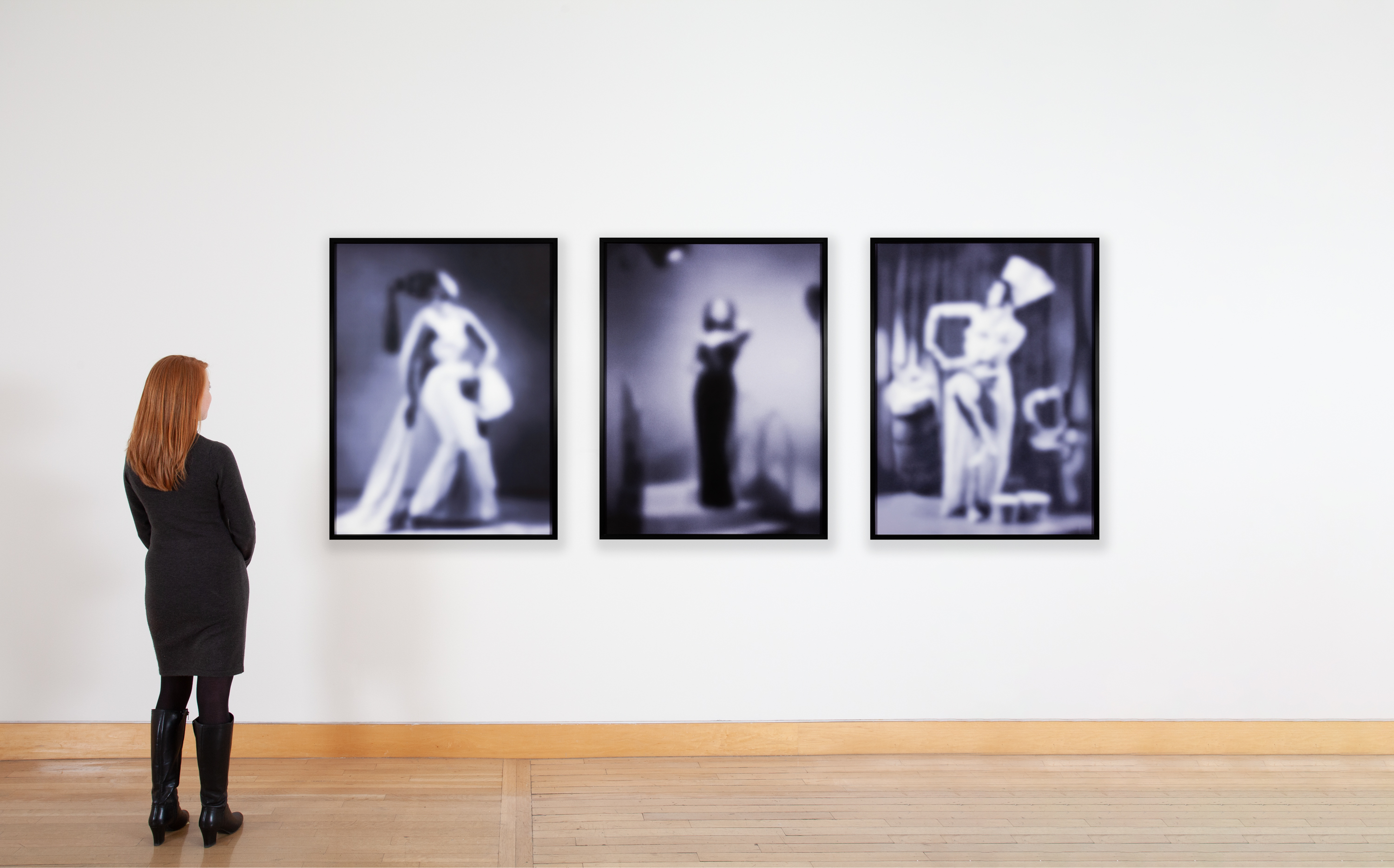 Color image of a figure looking at a triptych depicting three purple toned photographs of three performers out of focus framed in black on a white wall