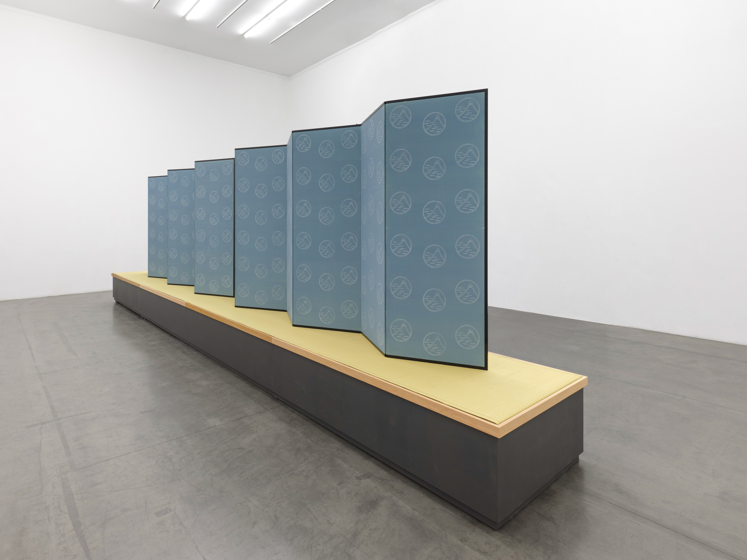 Installation view of a long blueish grey folding screen covered in woodblock prints of a mountain scene; the screen sits on a large pedestal