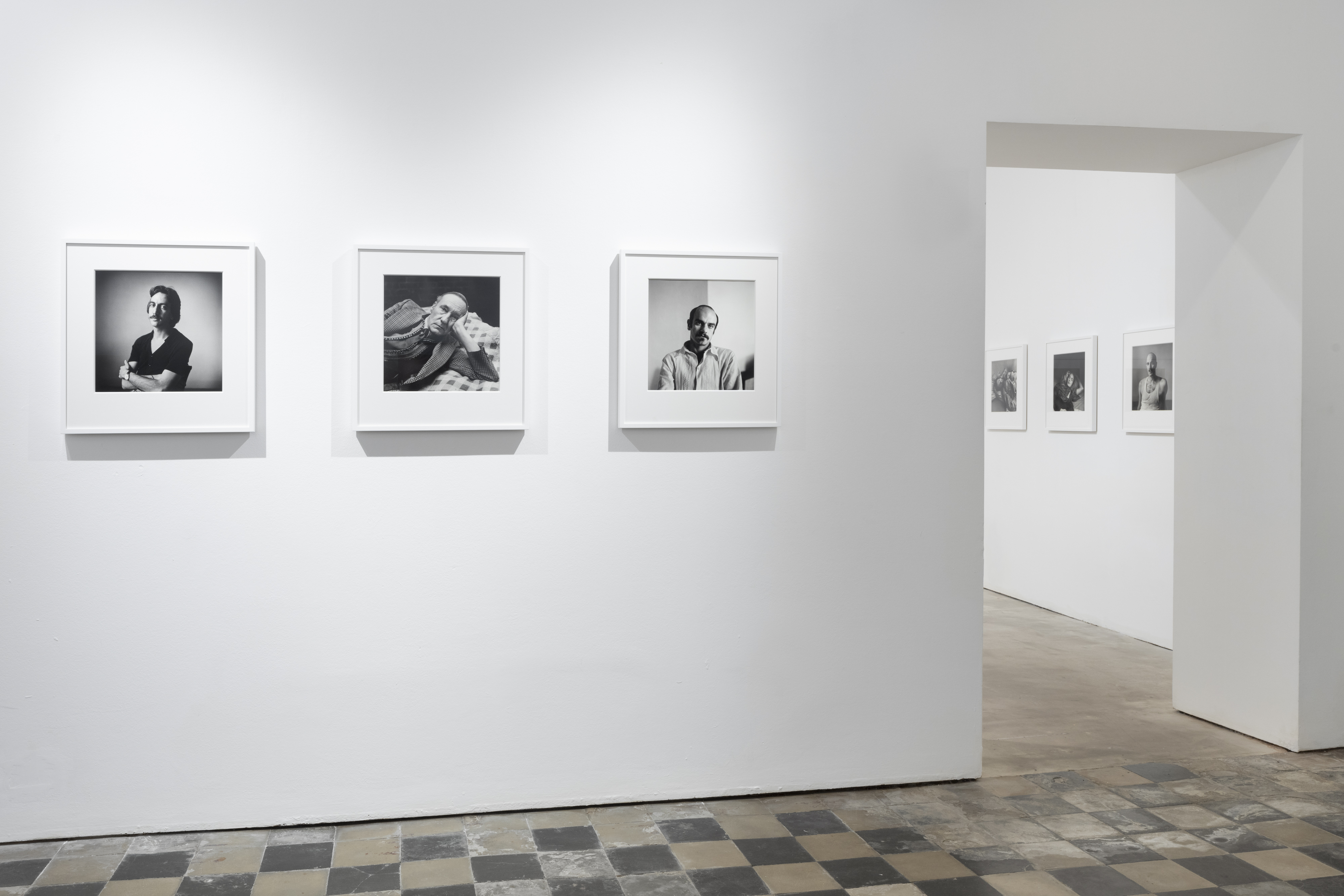 Color image of an exhibition of black and white photographs depicting various portraits framed in white on white walls