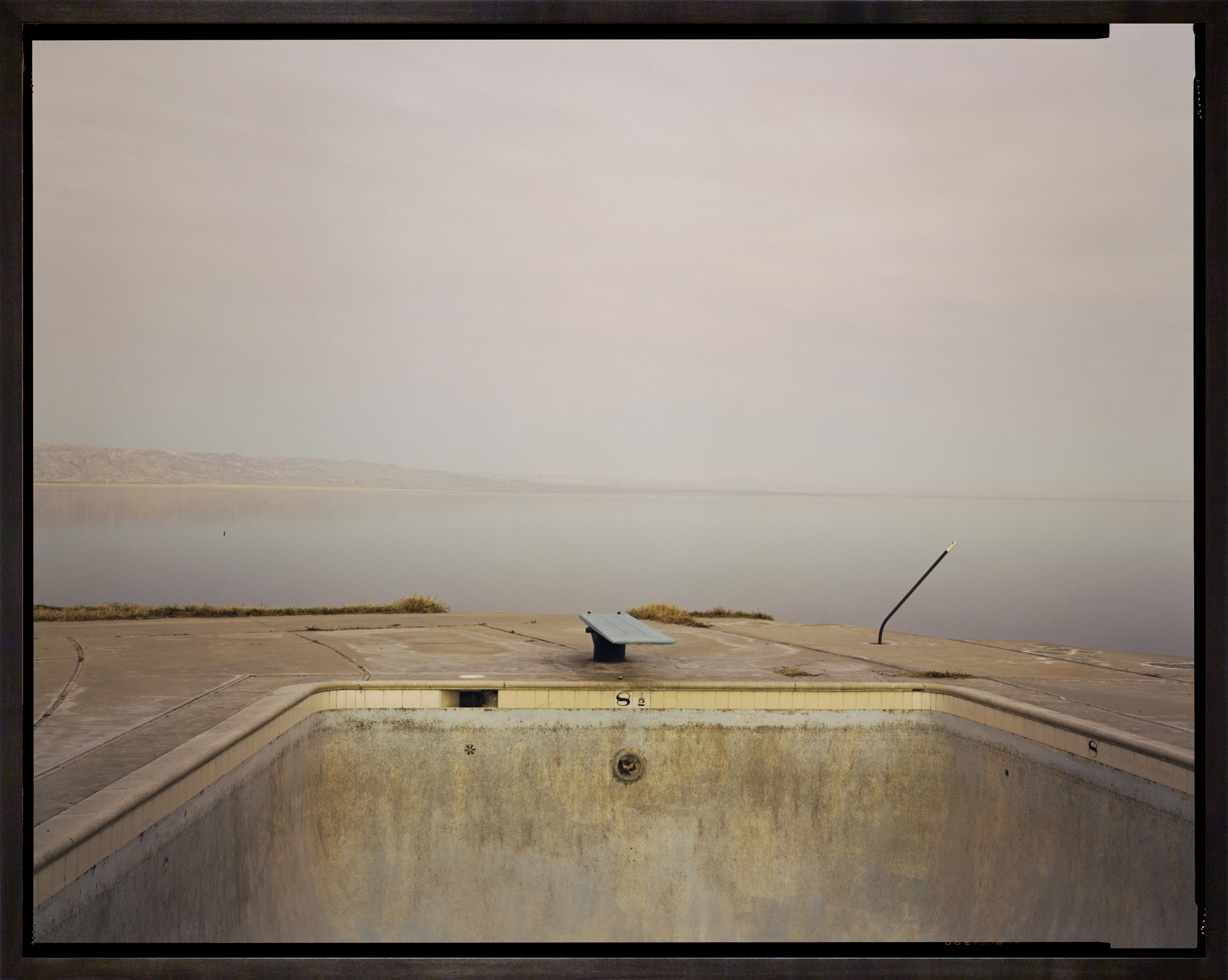 Color photograph of an abandoned swimming pool with a diving board alongside a view of the Salton Sea along the horizon framed in dark wood