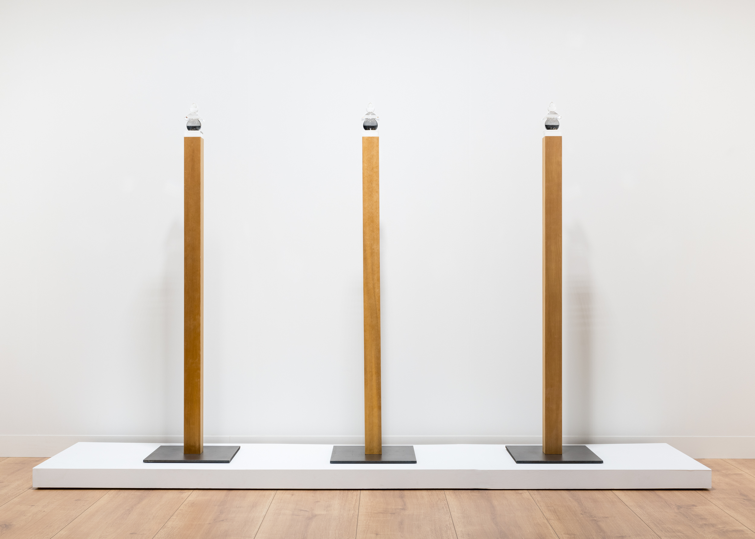 Color image of three sculptures compromised of wood, metal, and glass along a white wall