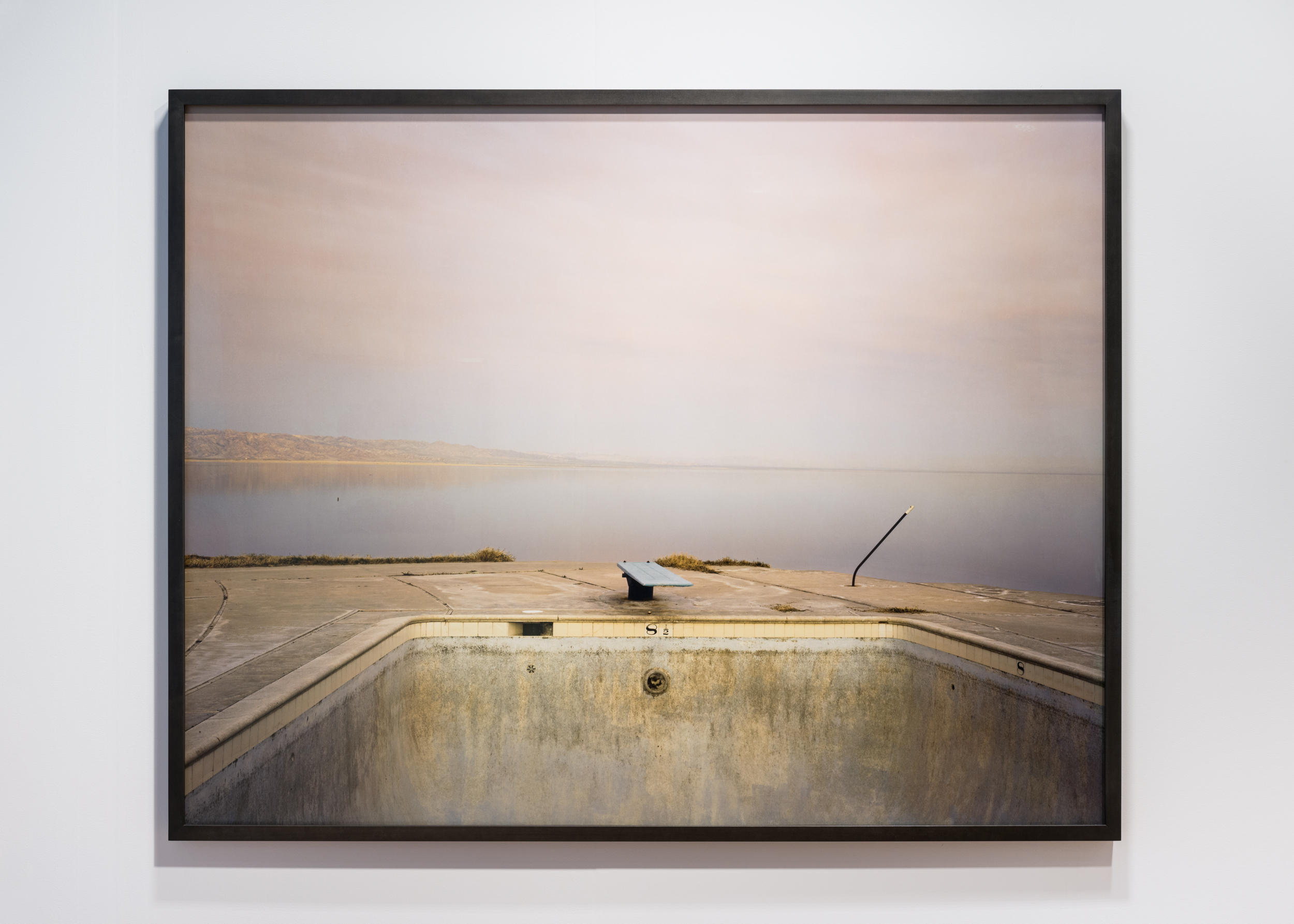 Color image of a color photograph depicting an empty swimming pool against a body of water framed in black on a white wall
